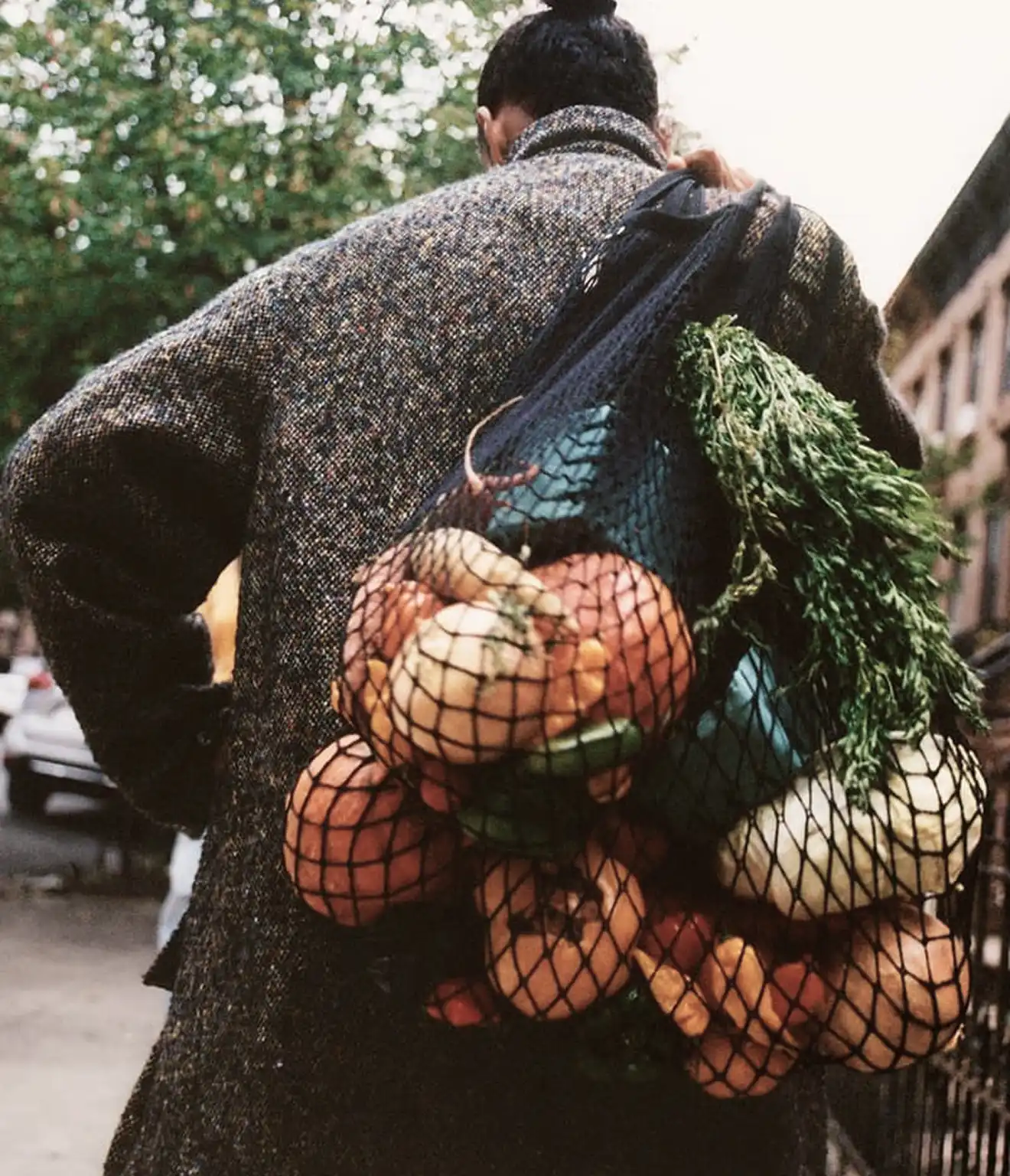 Person wearing a gray sweater carrying a mesh bag filled with various fresh vegetables on their shoulder.