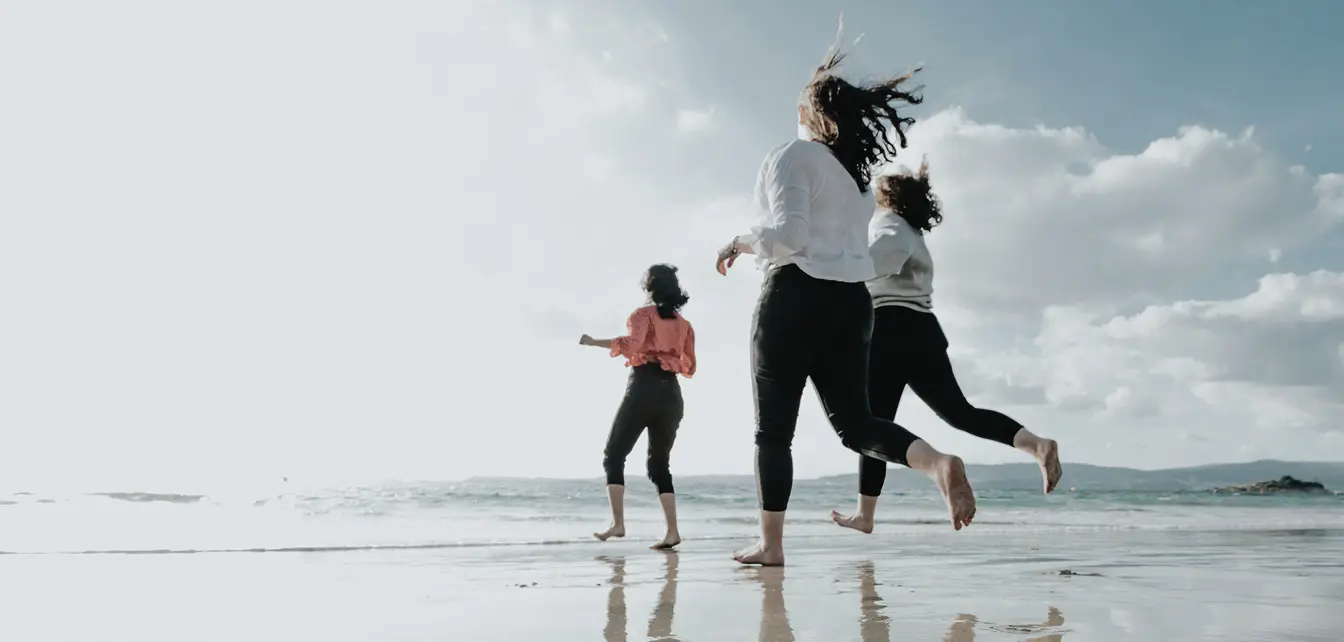 Three women joyfully running barefoot on a reflective beach under a cloudy sky.
