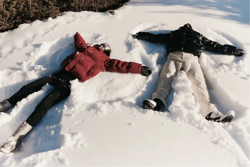 Two people lying on their backs in the snow making snow angels, wearing winter jackets and boots.