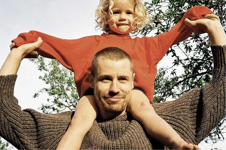 Smiling man wearing a brown sweater carrying a child in a red sweater on his shoulders outdoors with trees in the background.