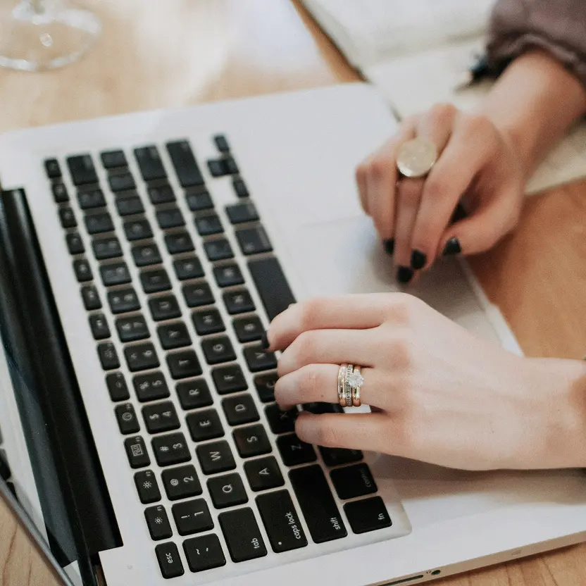 Close-up of hands typing on a laptop keyboard, with rings on fingers and black nail polish.