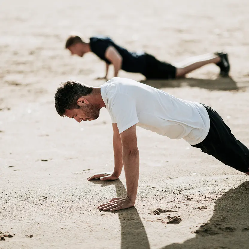 Two men doing push-ups on sandy ground in a workout session.