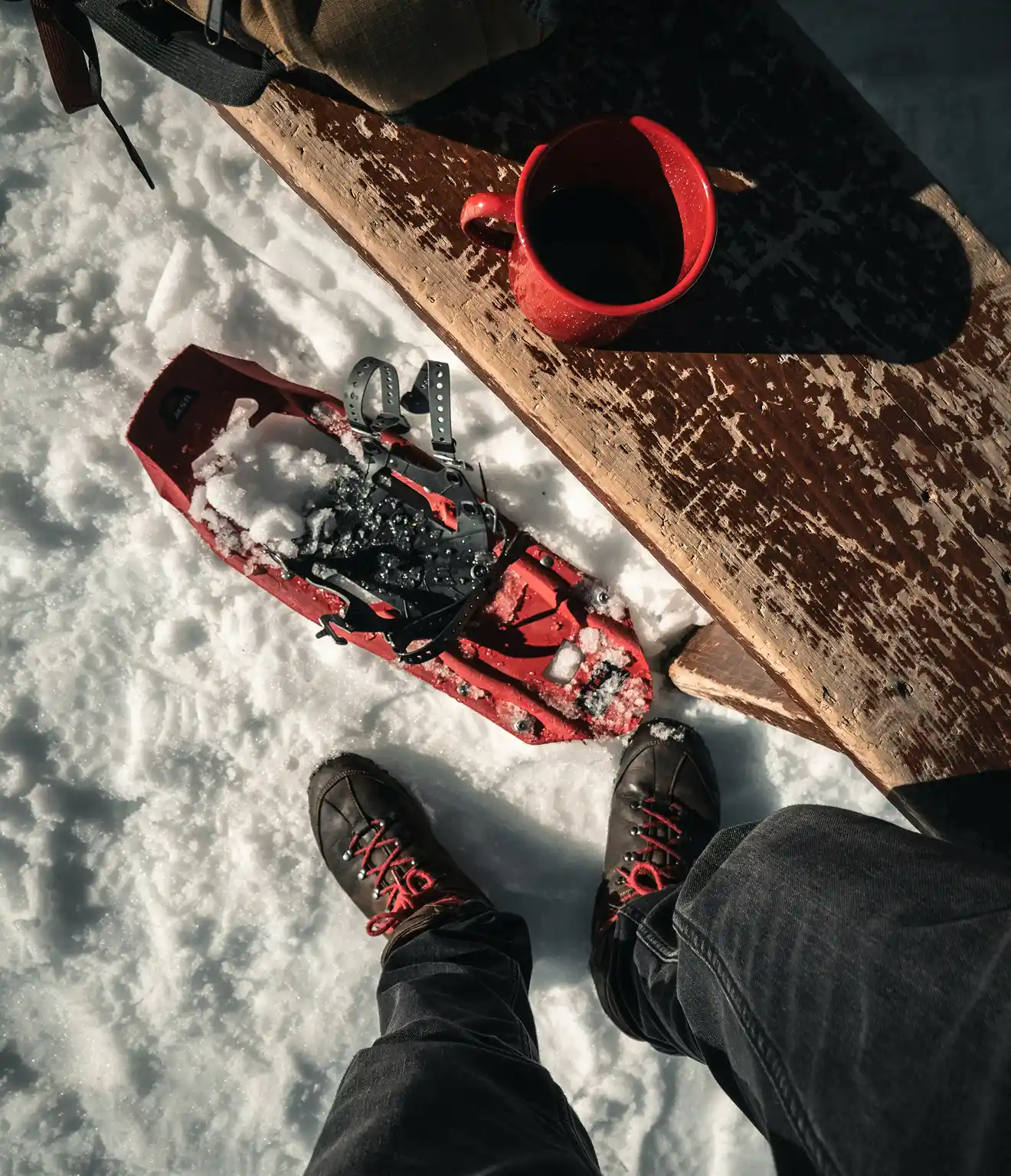 Person wearing black boots with red laces standing on snow next to a red snowshoe and a red mug on a wooden bench.