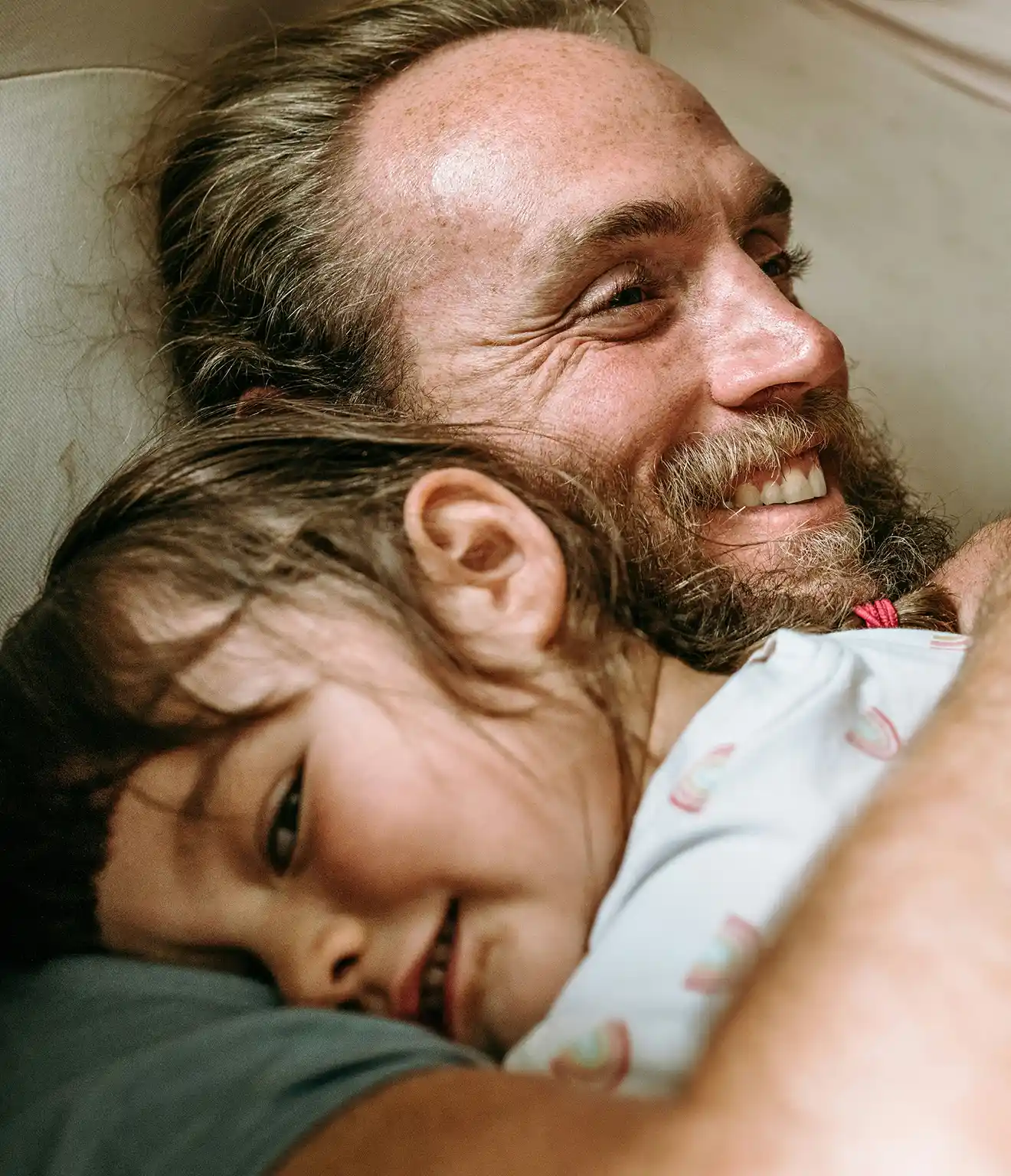 Close-up of a smiling bearded man hugging a young girl who rests her head on his shoulder.
