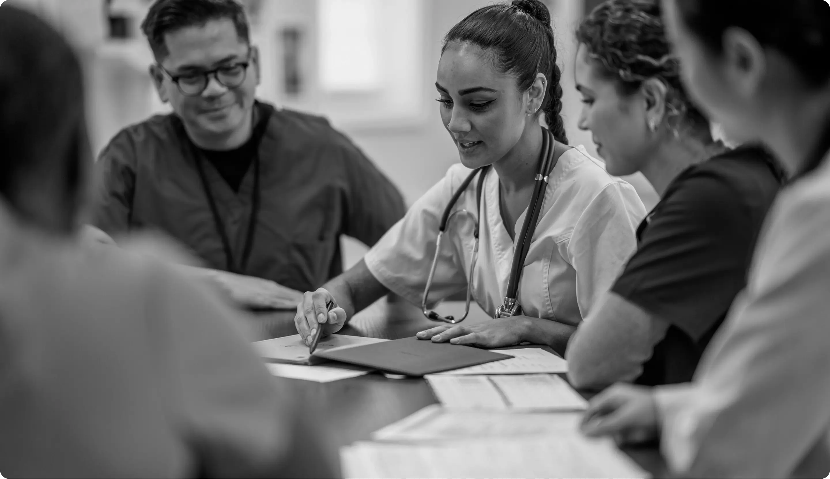 Medical team in discussion around a table with documents, featuring a female doctor with a stethoscope pointing at papers.