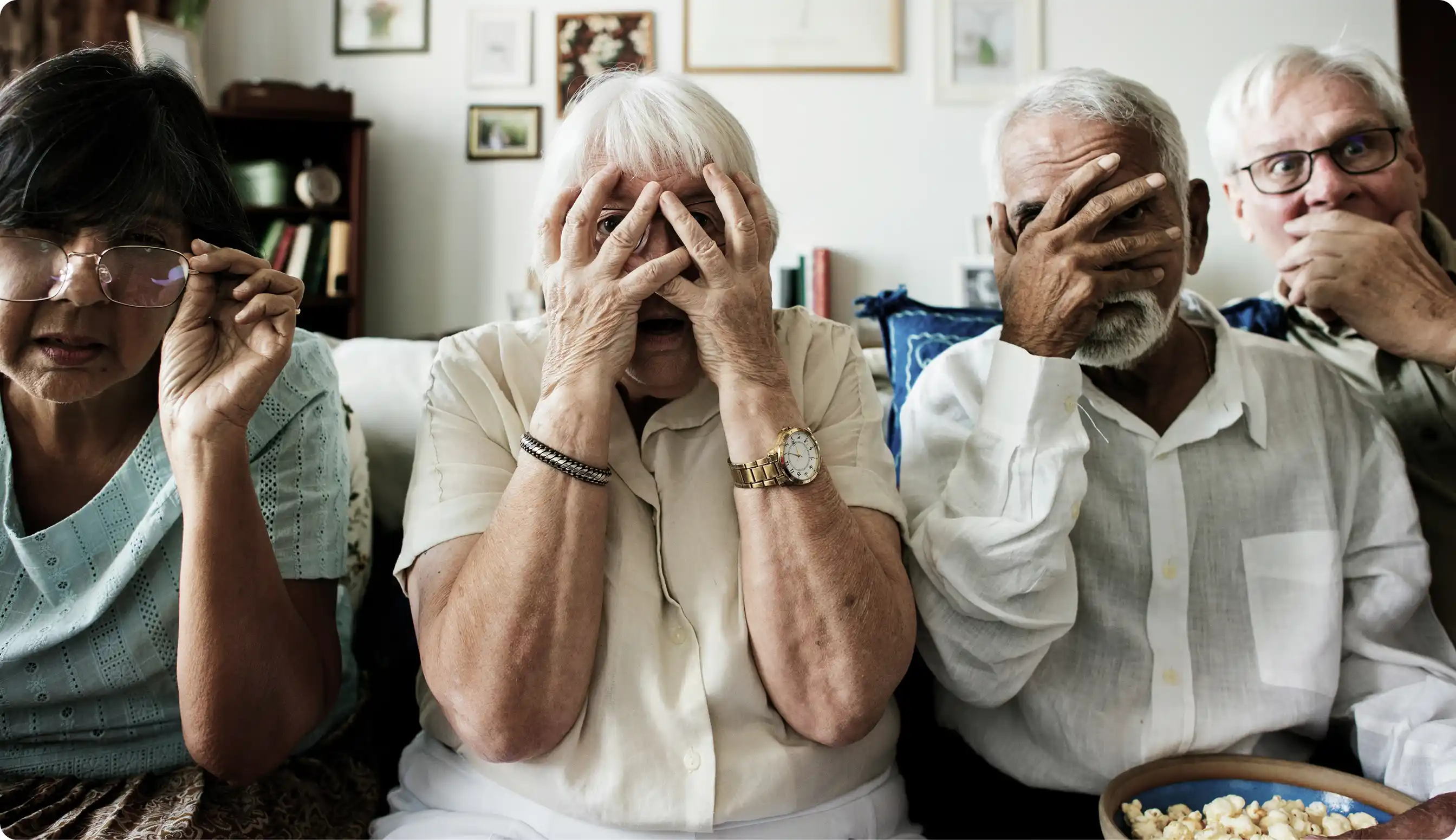 Four elderly people sitting together on a couch with playful expressions, covering or partially covering their faces with their hands.