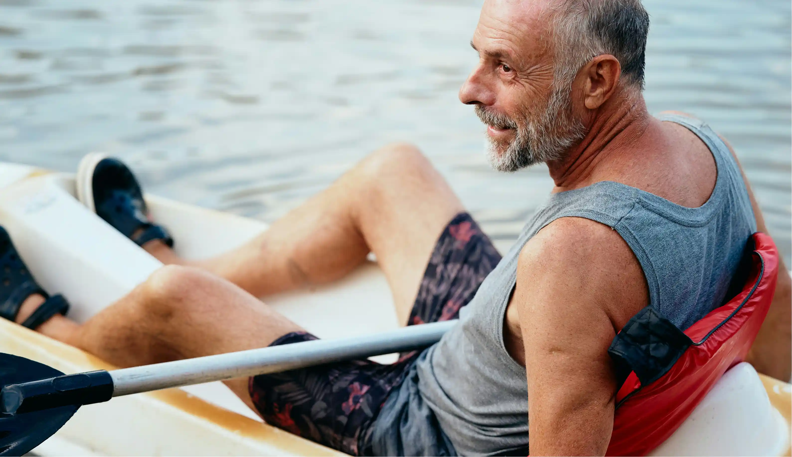 Mature man with gray beard sitting in a kayak on calm water holding a paddle.