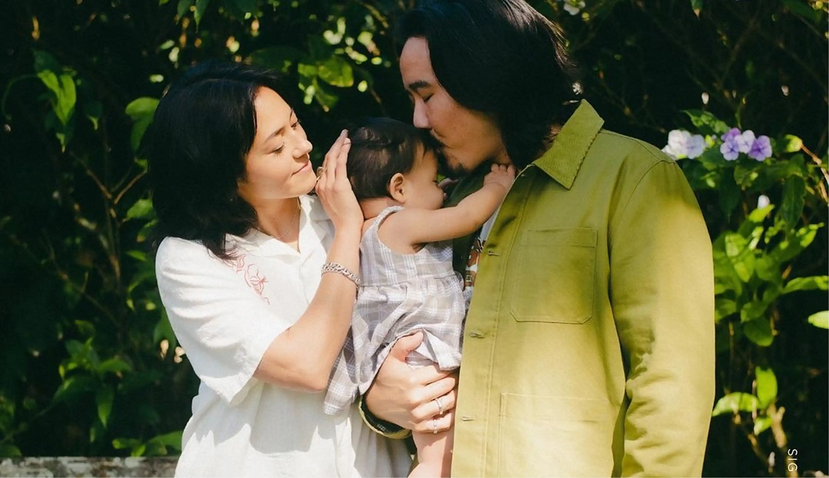 Man kissing the forehead of a baby wearing a checkered dress while a woman gently touches the baby's head outdoors.
