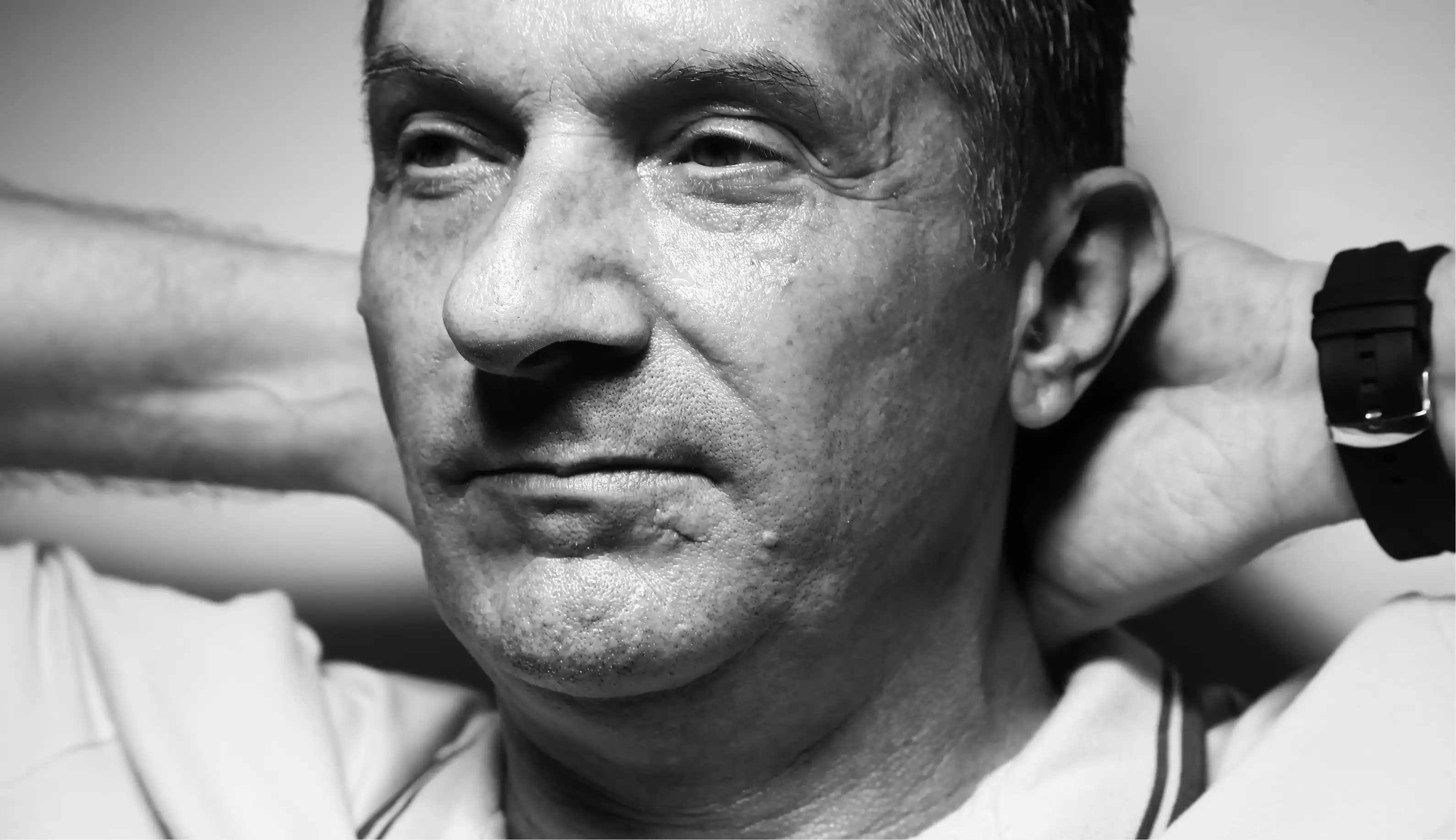 Black and white close-up portrait of a mature man with his hands behind his head, wearing a watch and a collared shirt.