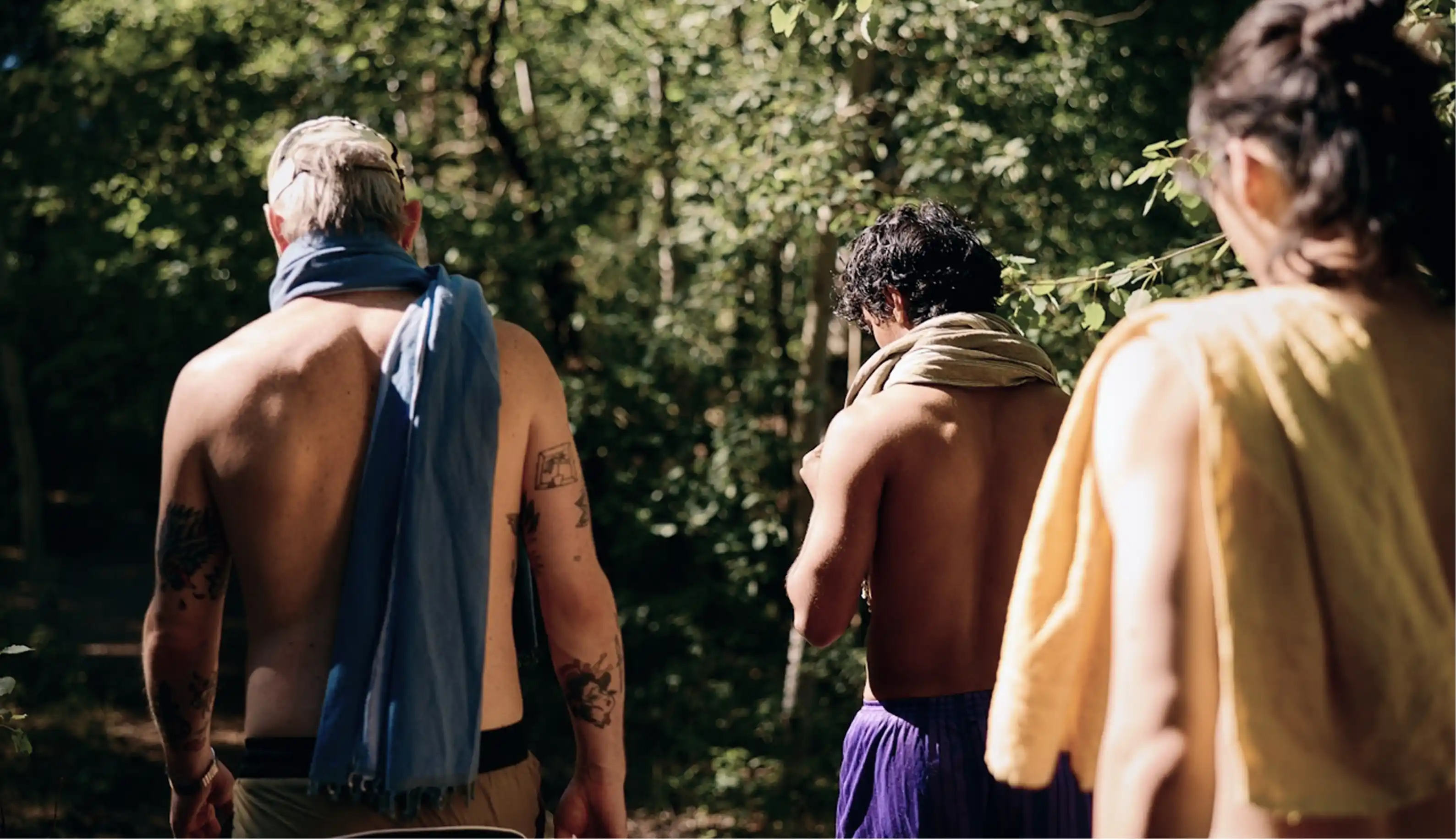 Three shirtless people walking outdoors with towels draped around their necks, surrounded by green trees.
