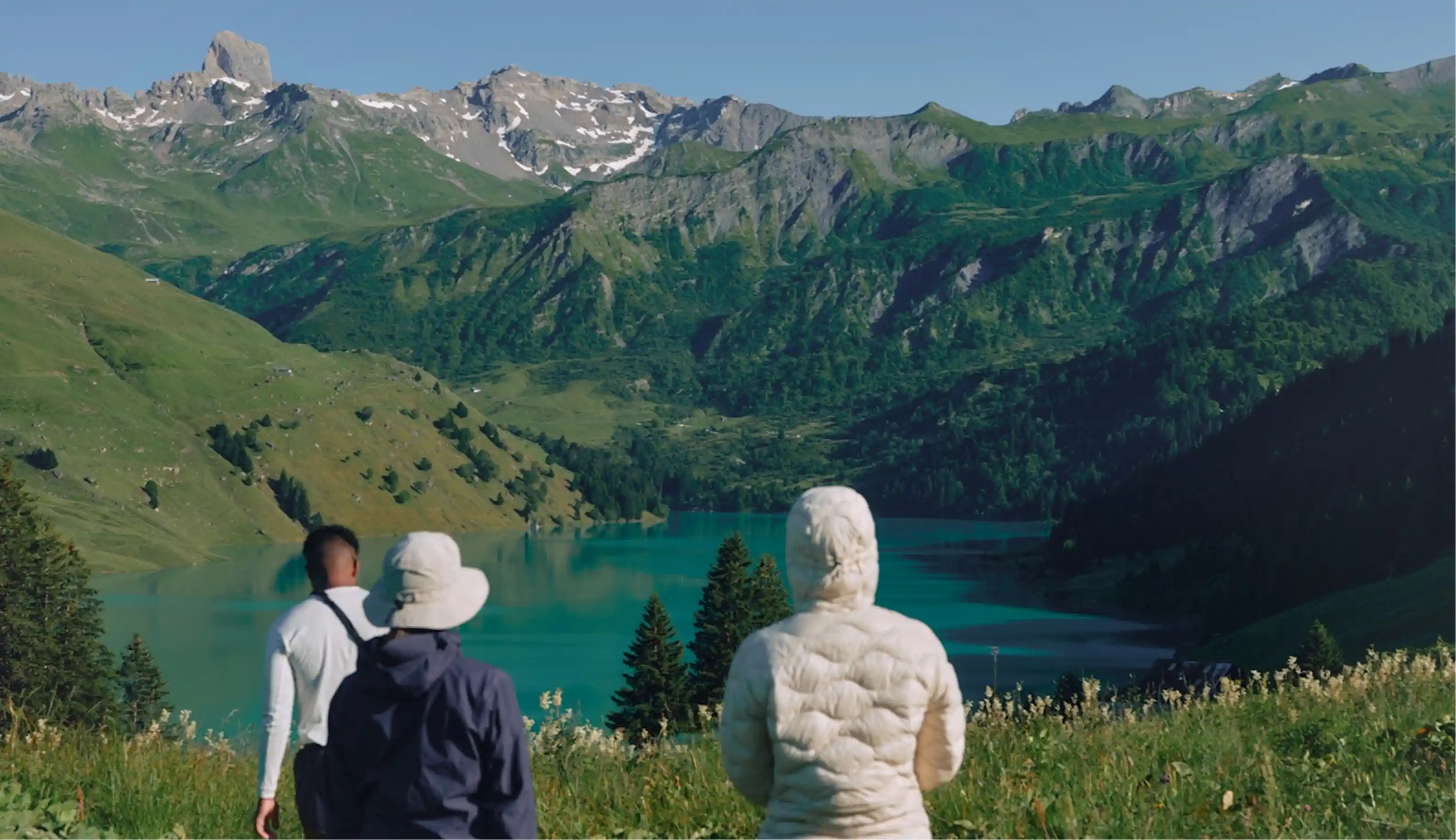 Three hikers standing on a grassy hill overlooking a turquoise lake surrounded by green mountains with patches of snow under a clear blue sky.