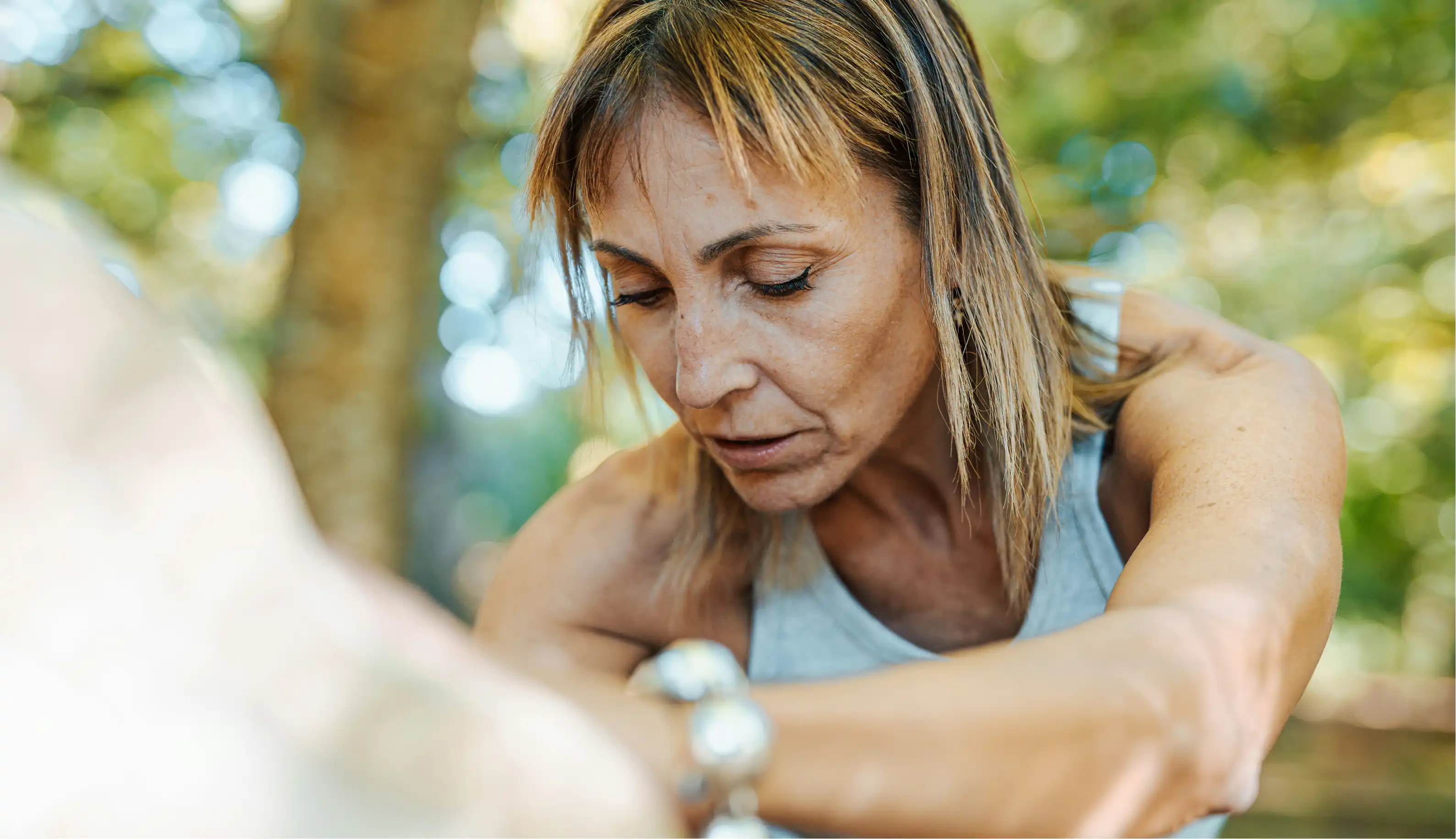 Focused middle-aged woman stretching her leg outdoors with a blurred natural background.
