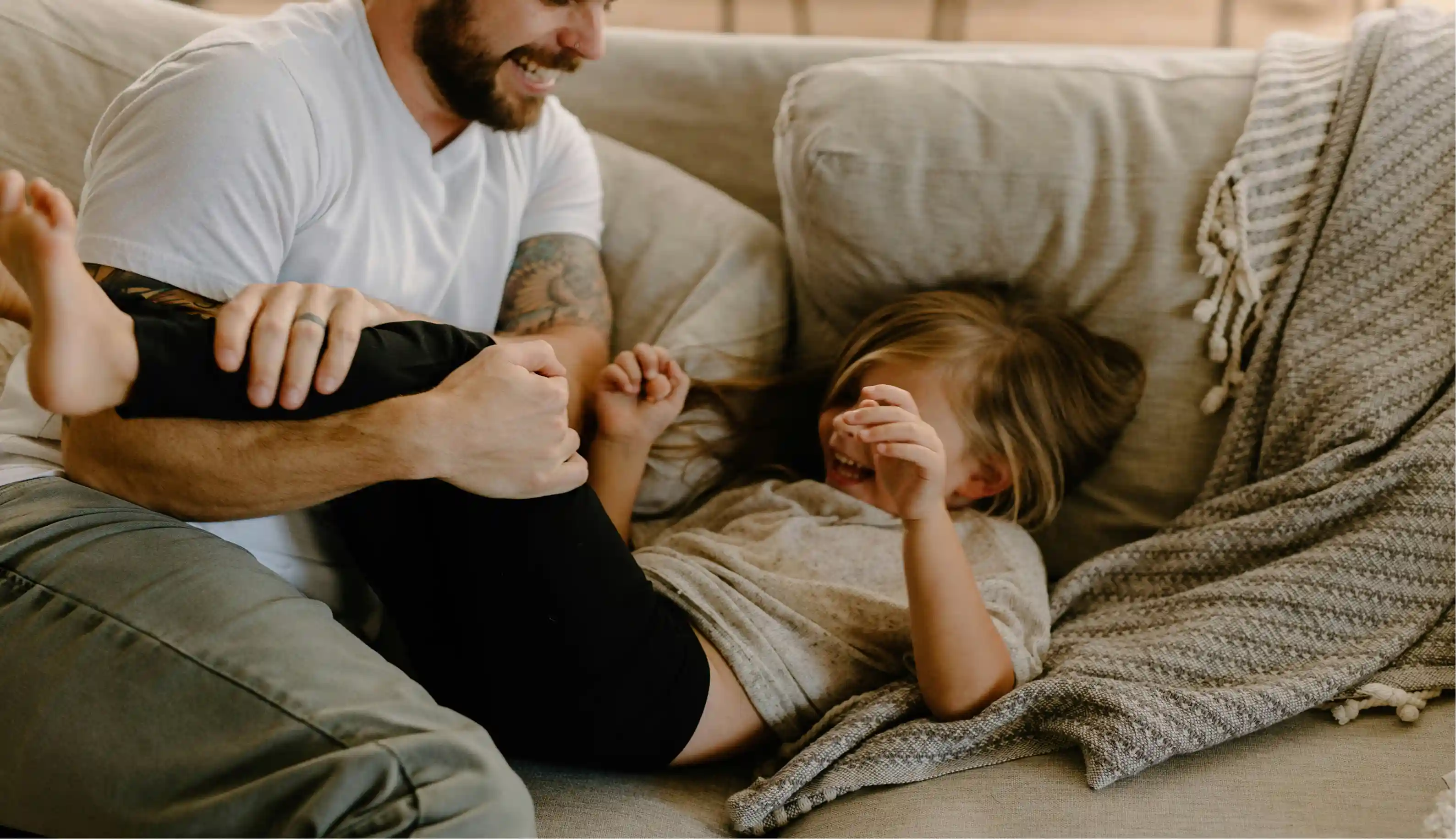 Father playfully tickling his laughing young child on a beige couch with a knitted blanket.