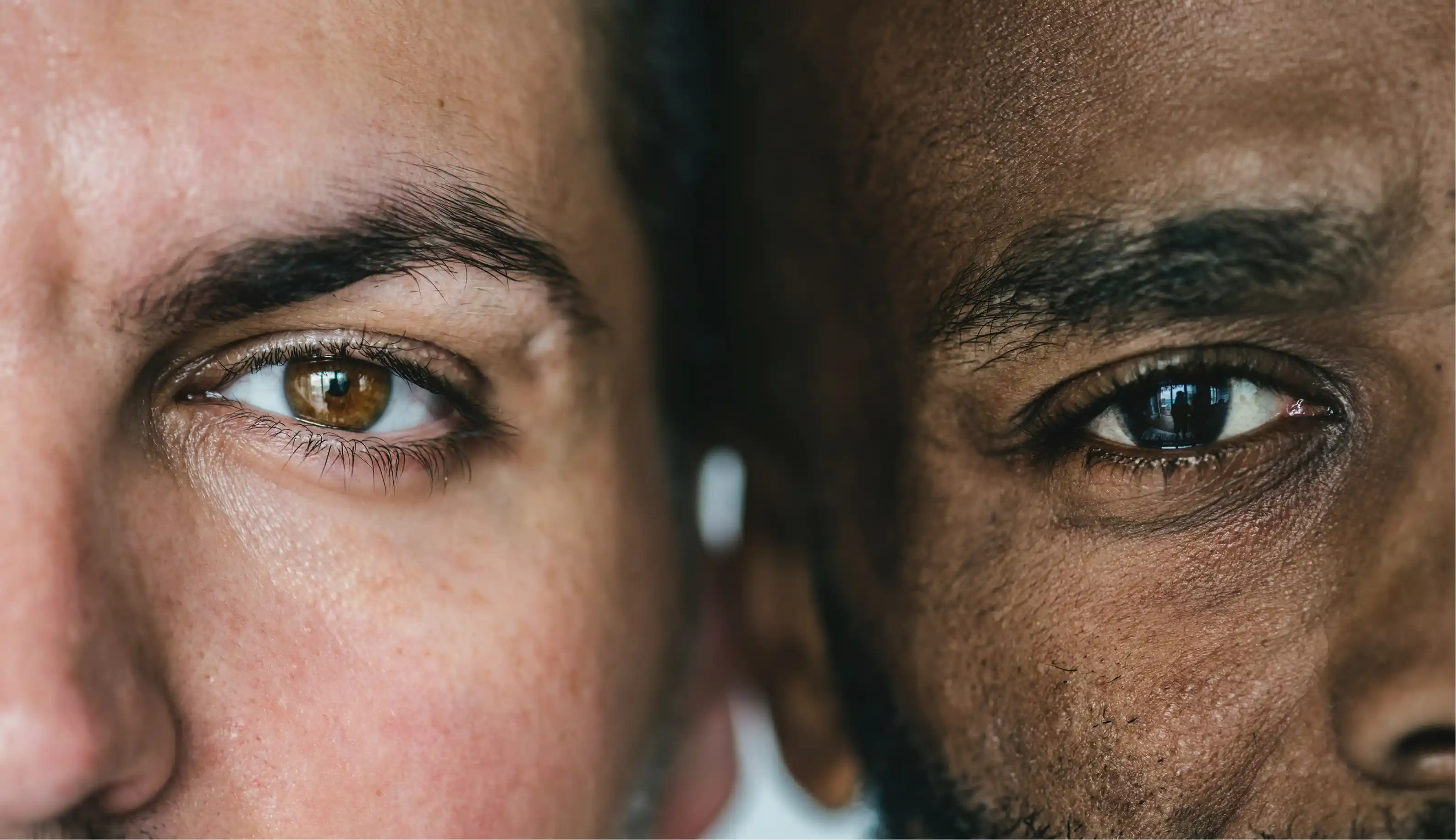 Close-up of two men's faces side by side, focusing on their eyes with one having brown eyes and the other dark brown eyes.