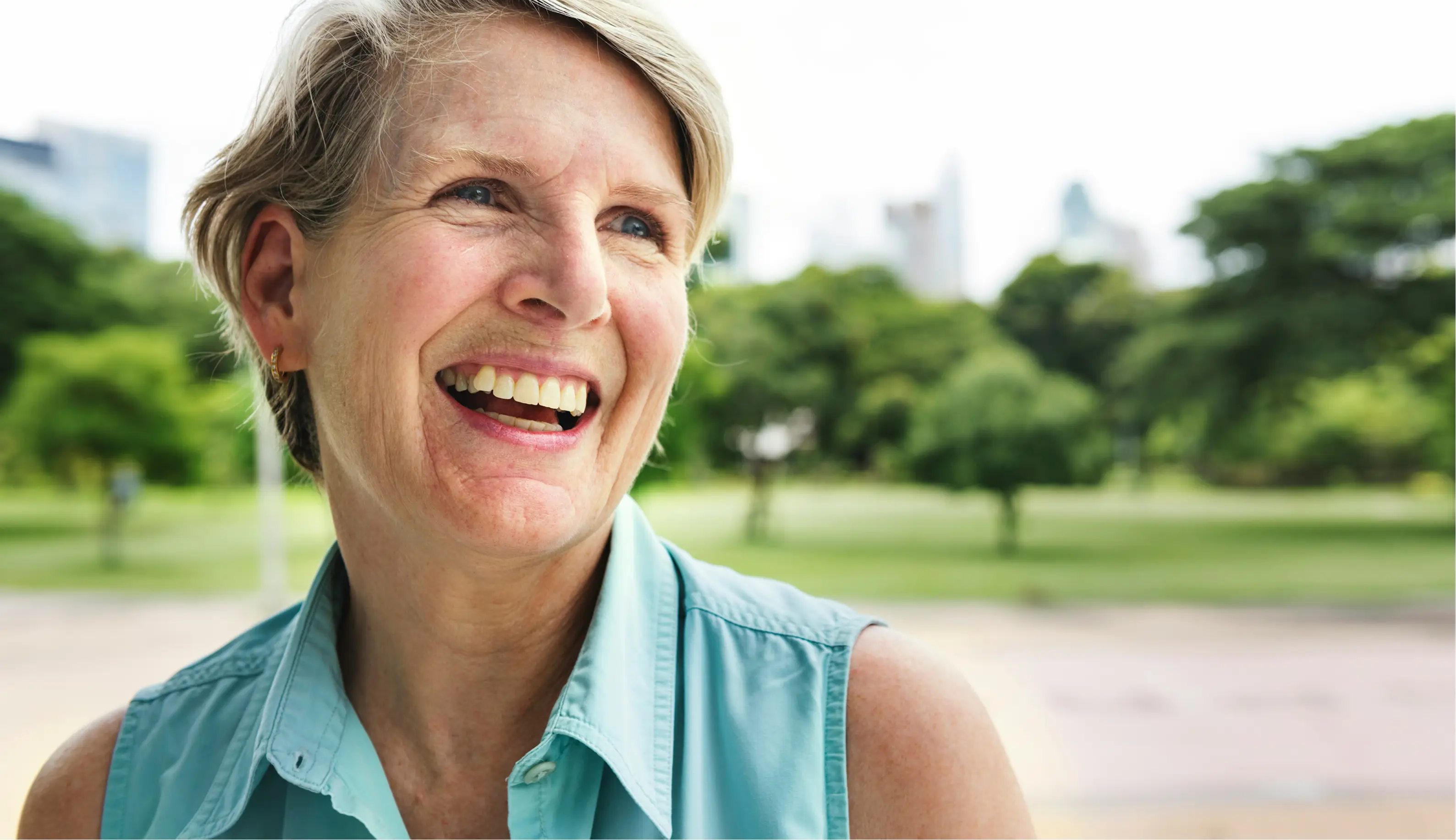 Smiling mature woman with short hair wearing a sleeveless light blue shirt outdoors in a park.