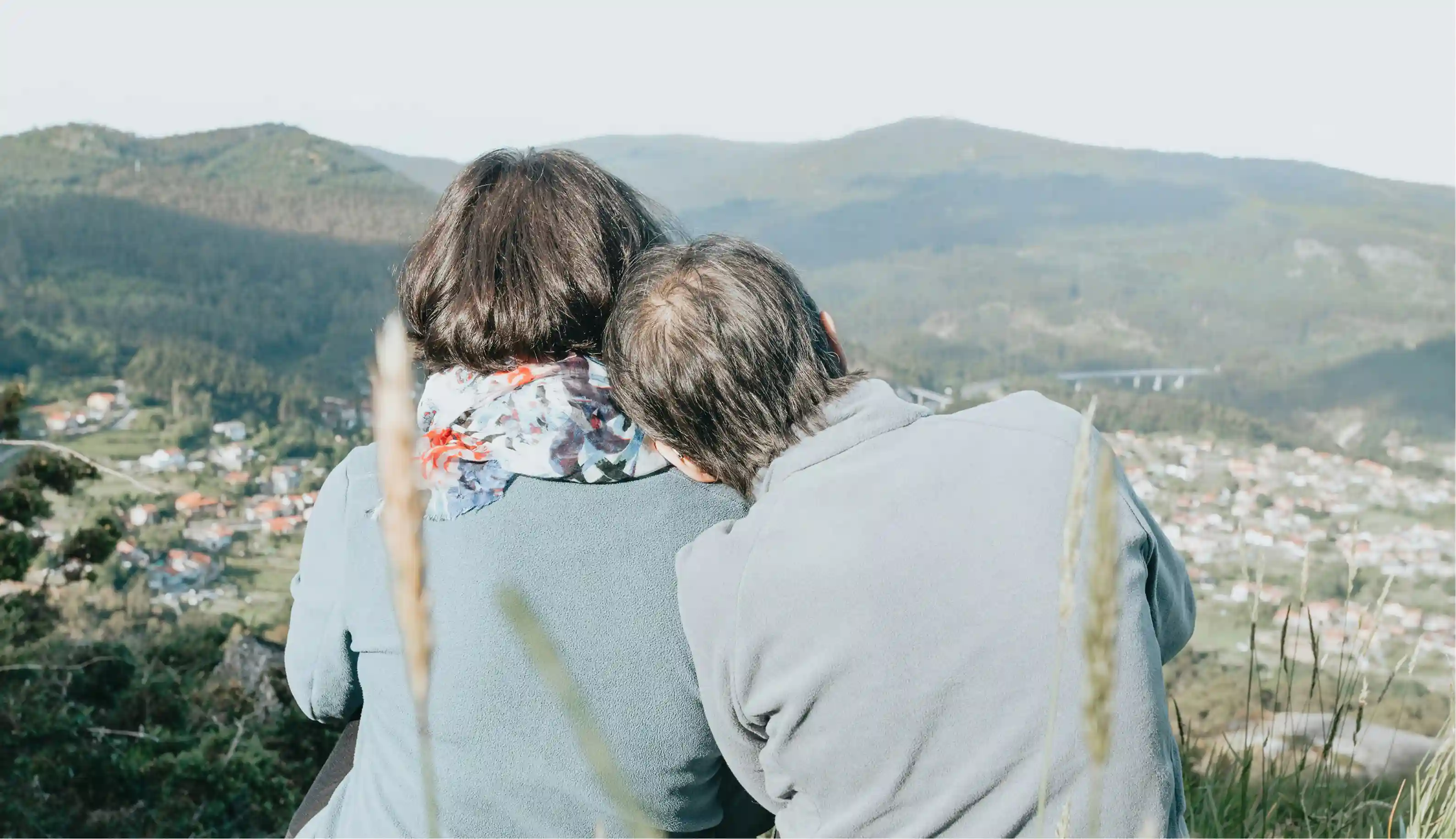 Older couple sitting outdoors on a hill, with the man resting his head on the woman's shoulder, overlooking a valley with houses and mountains.
