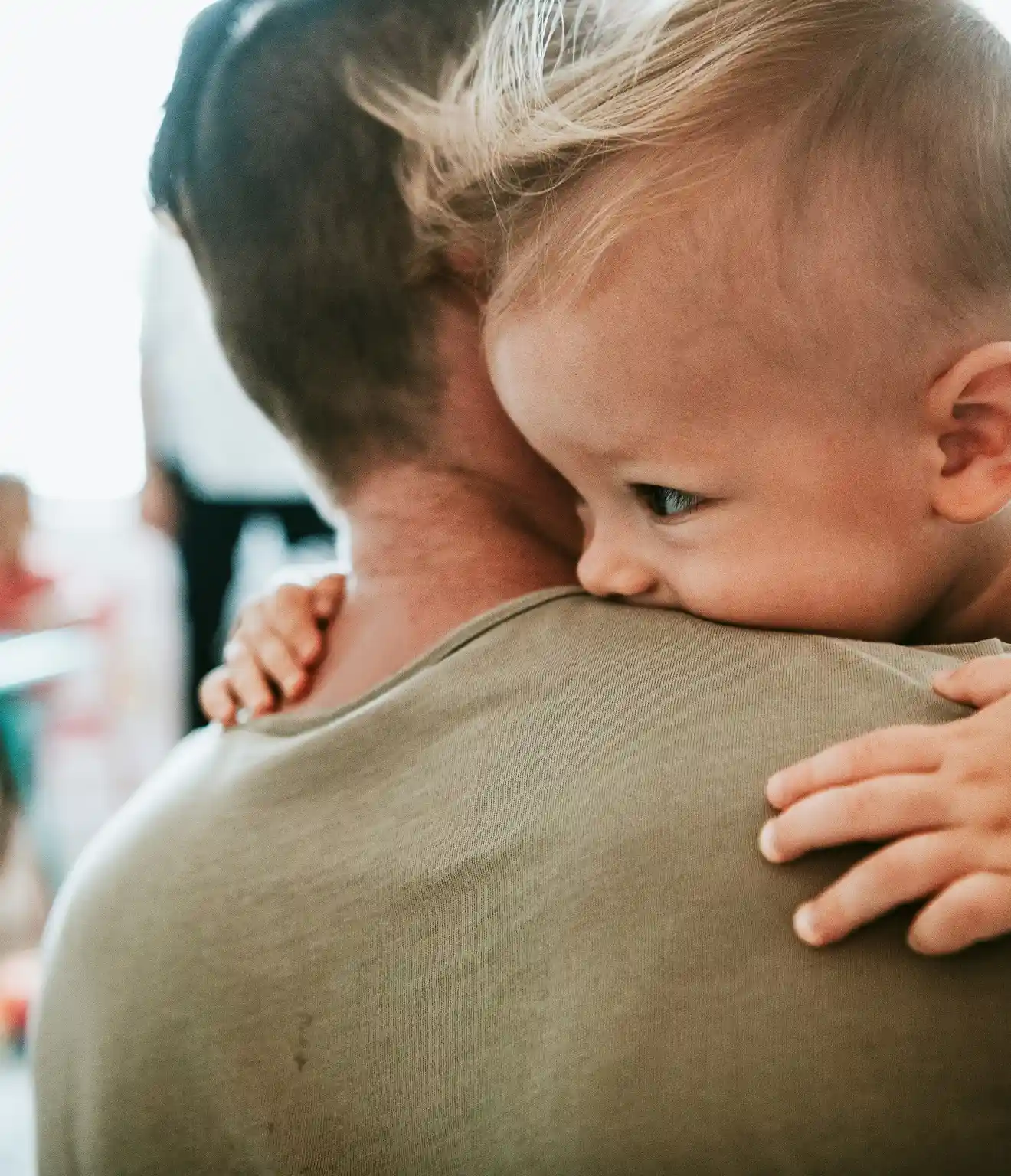 Toddler with blonde hair resting on an adult's shoulder, holding on gently.