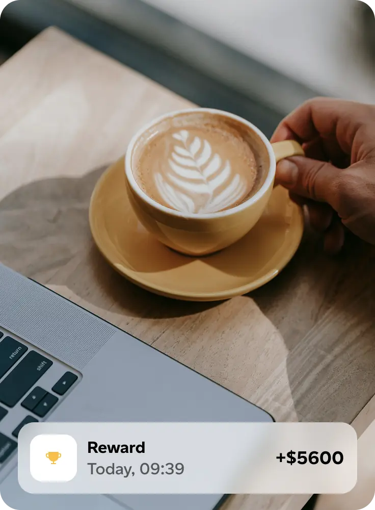 Hand holding a yellow cup of latte with leaf latte art on a wooden table next to a laptop, with an on-screen notification showing a reward of +$5600.