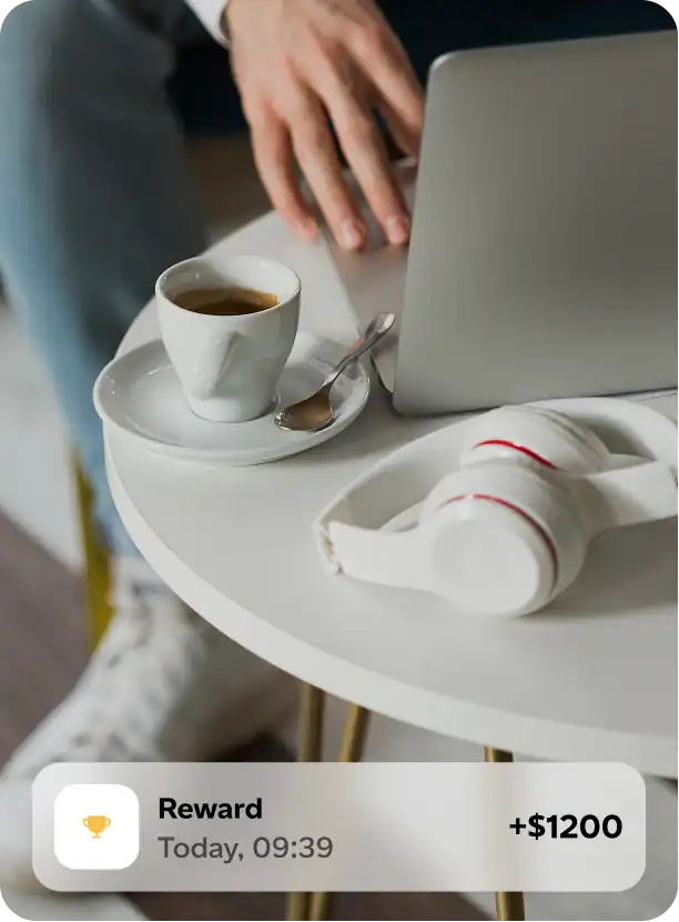 Person using a laptop at a white round table with a cup of coffee and white headphones resting on the table, with an on-screen notification showing a $1200 reward.