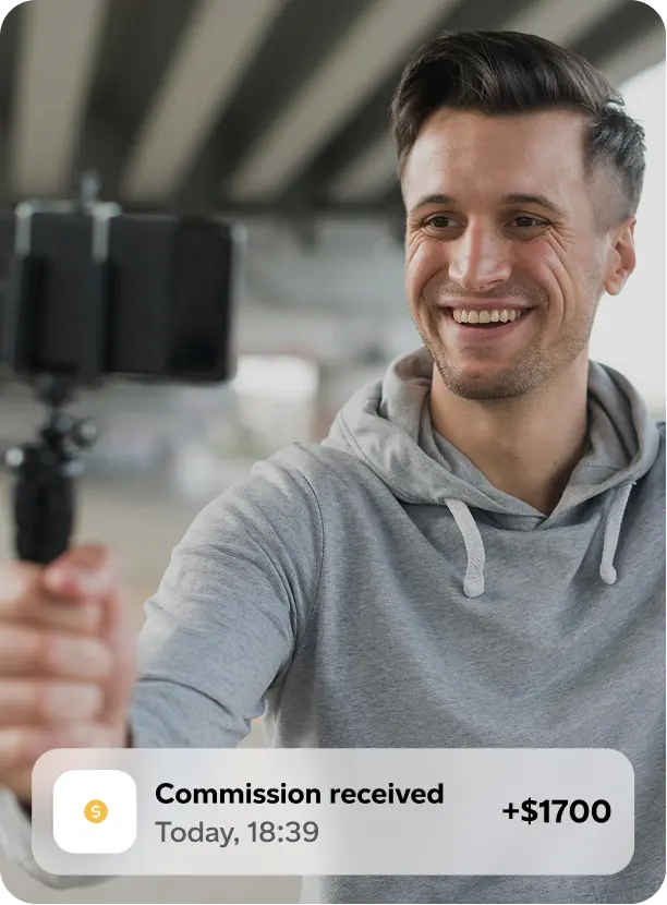 Smiling man in gray hoodie holding a smartphone on a selfie stick with an overlay notification showing a commission received of $1700.