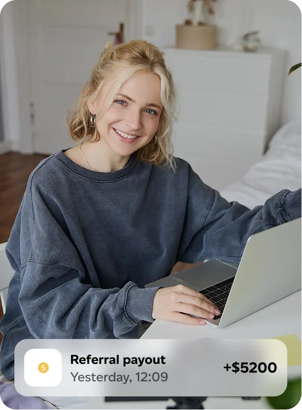 Smiling blonde woman in a dark gray sweatshirt sitting at a desk with a laptop, with a notification showing a referral payout of $5200.