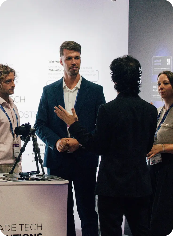 Four professionals engaged in conversation at a trade show booth, with a camera on a tripod displayed on the counter.