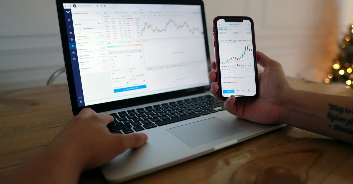 Trader using laptop and smartphone simultaneously to monitor stock charts — prop trading strategies