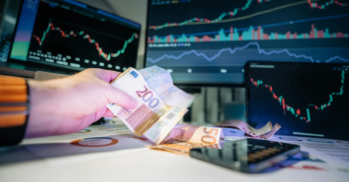 Hand holding euro banknotes in front of multiple screens showing forex charts — how to get a funded trading account