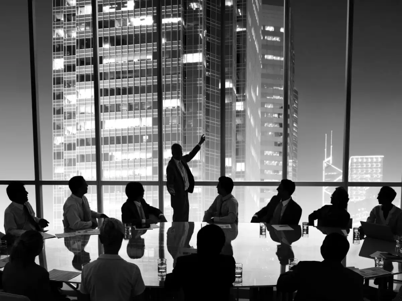 Silhouettes of businesspeople in a meeting room with a presenter pointing to a large cityscape through floor-to-ceiling windows.