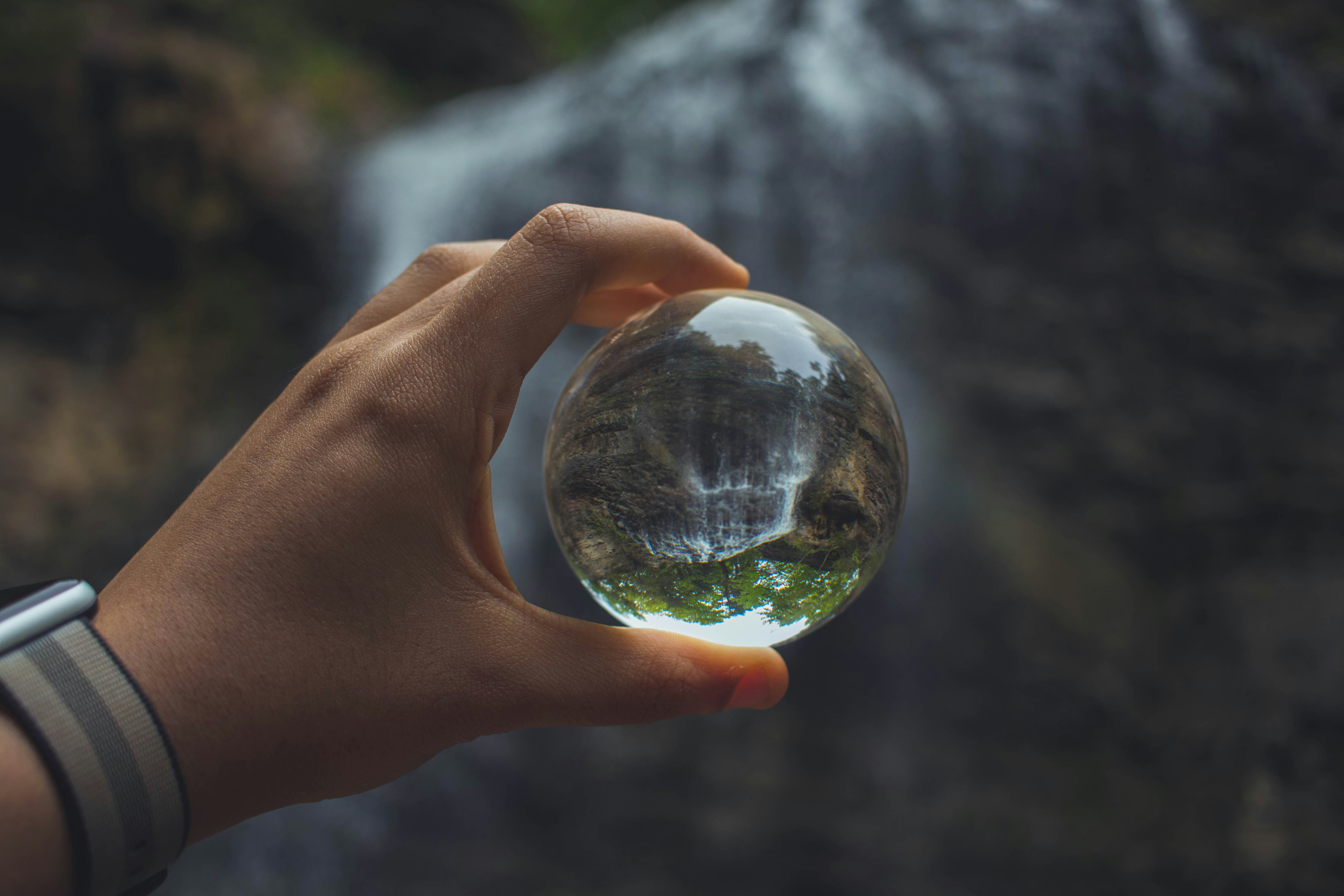 Hand holding a clear glass sphere that reflects an inverted image of a waterfall and surrounding greenery.