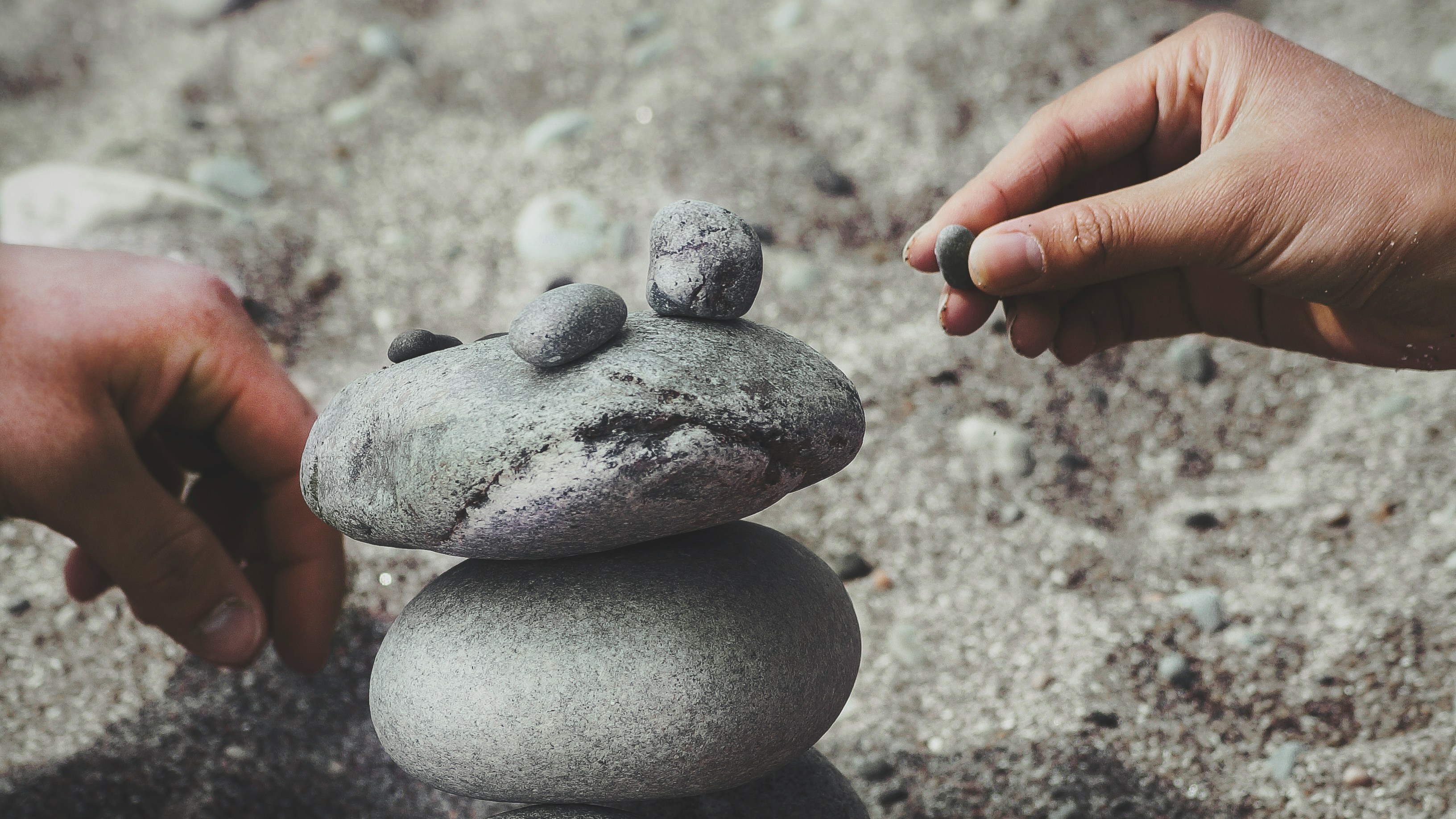 Two hands carefully stacking small and medium gray stones on a sandy surface.