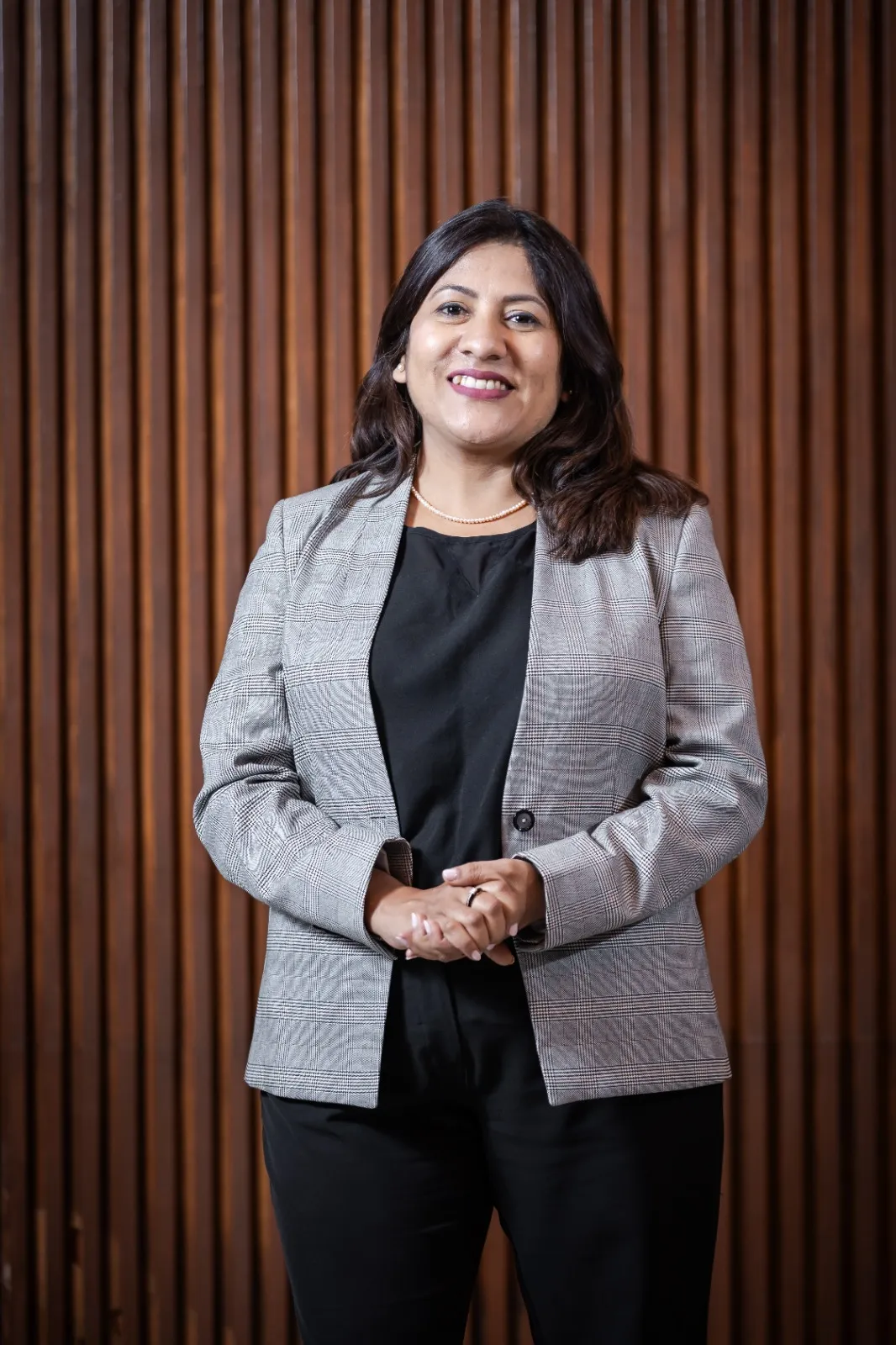 Smiling woman wearing a grey plaid blazer and black outfit standing in front of a wooden slat wall.