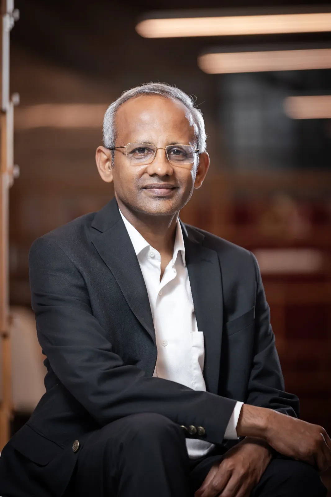 Portrait of a middle-aged man with glasses wearing a black suit and white shirt, sitting with hands folded, smiling slightly.