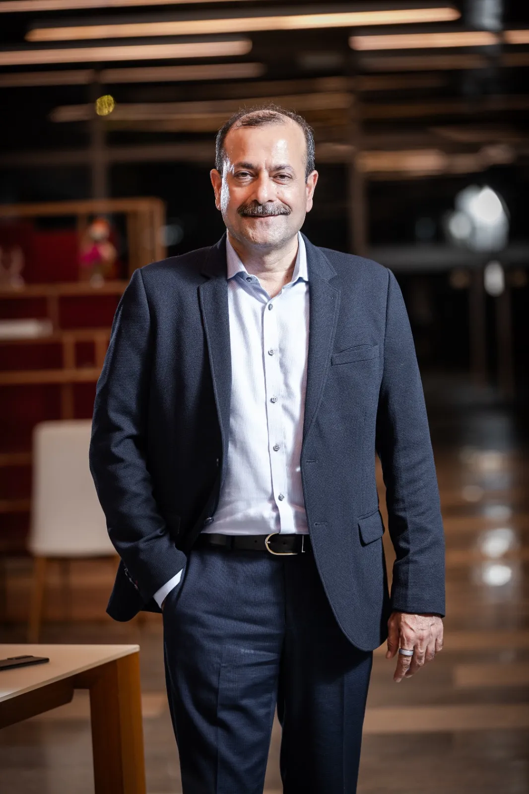 Smiling middle-aged man in a dark suit and light blue shirt standing indoors with one hand in his pocket.