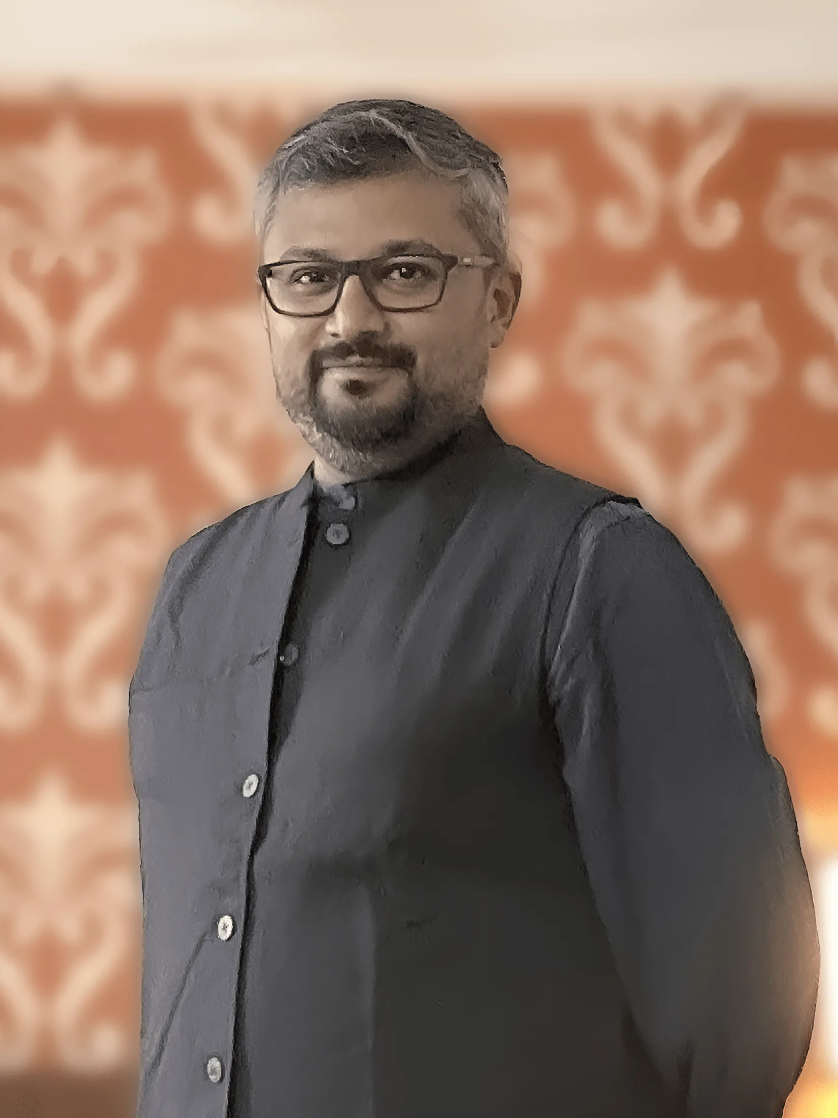 Man with short gray hair and glasses wearing a dark buttoned-up vest, standing in front of an orange patterned background.