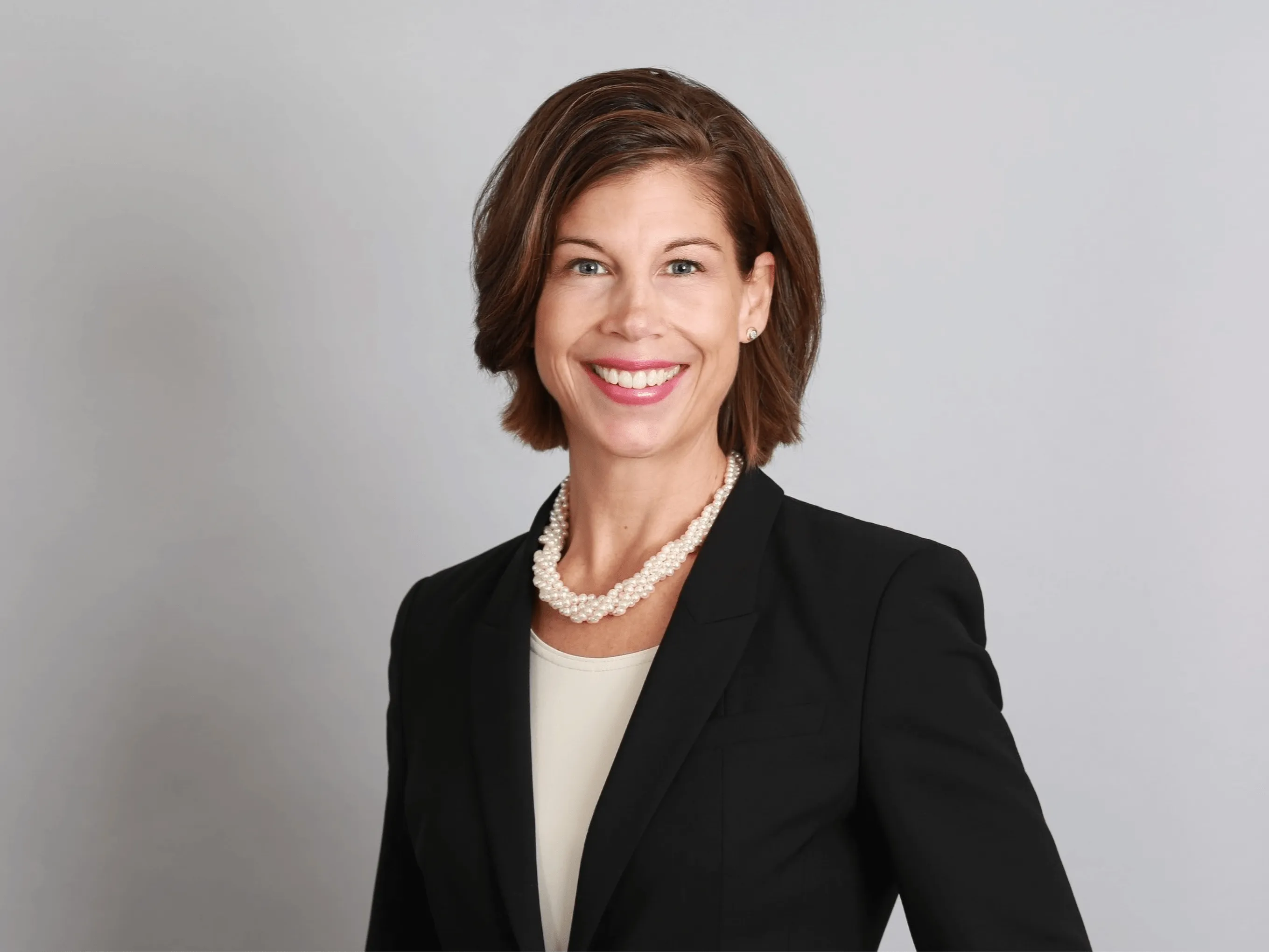 Smiling woman with short brown hair wearing a black blazer, white blouse, and pearl necklace against a light gray background.