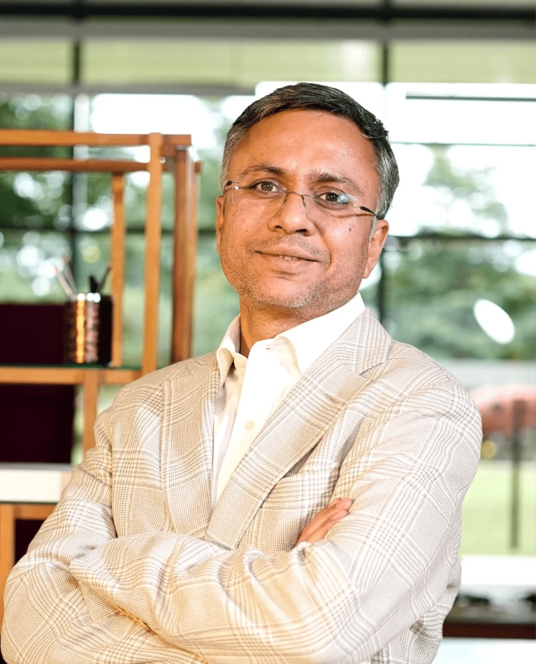 Middle-aged man wearing glasses and a light plaid blazer standing with arms crossed in a modern office setting.