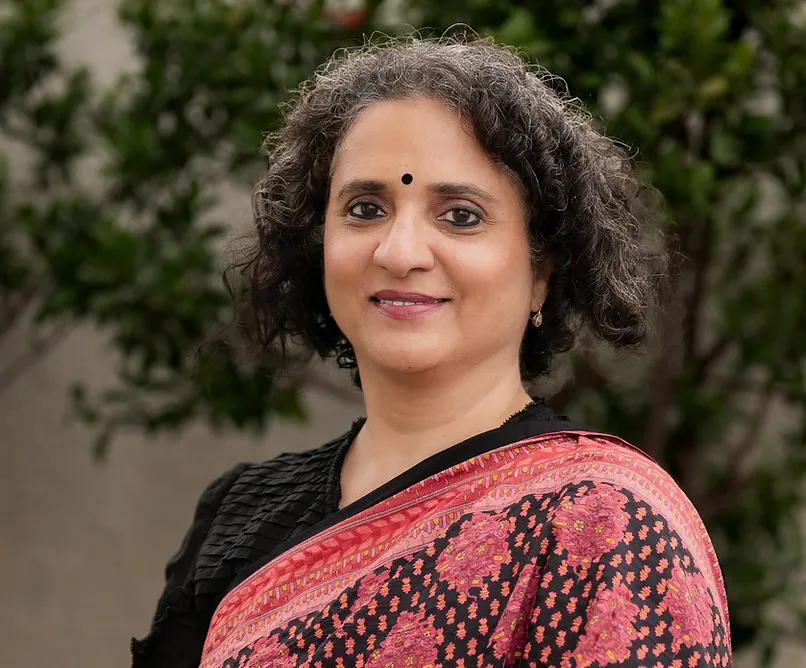 Smiling woman with curly hair and a bindi wearing a black and pink floral-patterned sari, standing outdoors with greenery in the background.