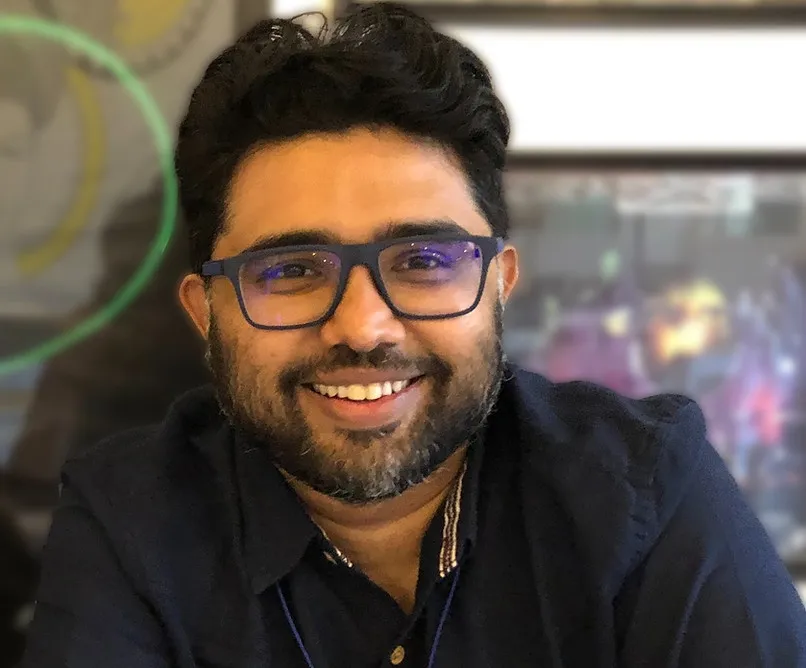 Smiling bearded man wearing glasses and a dark shirt, sitting indoors with blurred background.