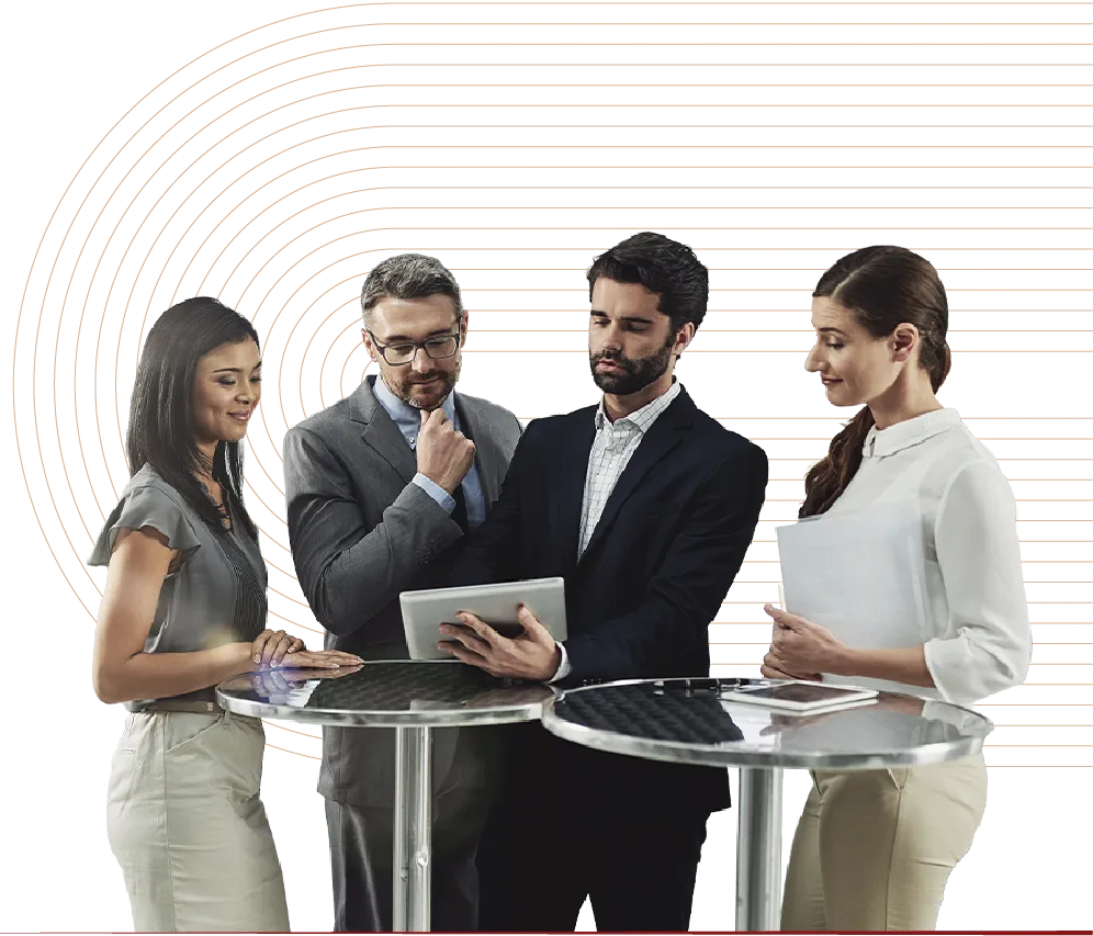 Four business professionals standing around two tables reviewing information on a tablet device.
