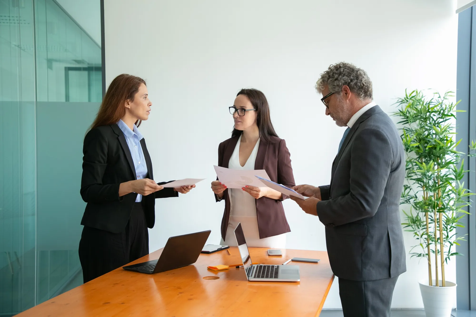 Three business professionals standing around a table with laptops and papers in a modern office.