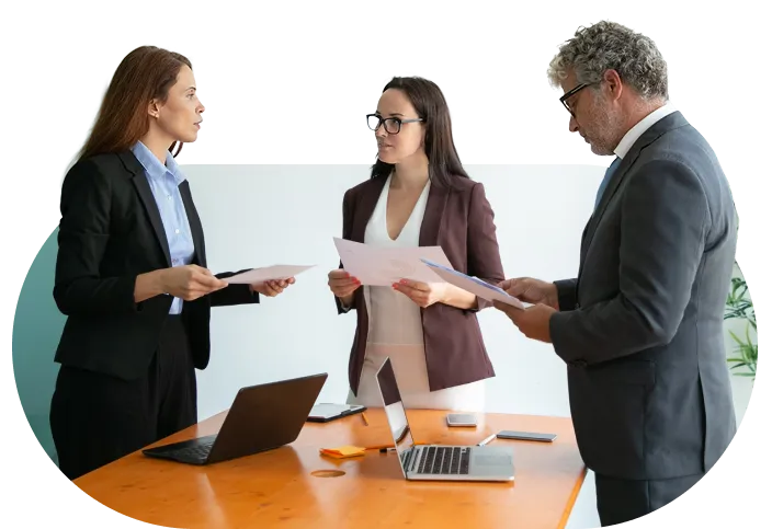 Three business professionals discussing documents around a table with laptops and phones.