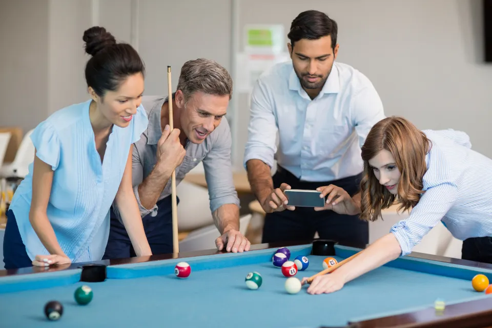 Four coworkers playing pool in an office, one aiming a shot while another takes a photo with a smartphone.