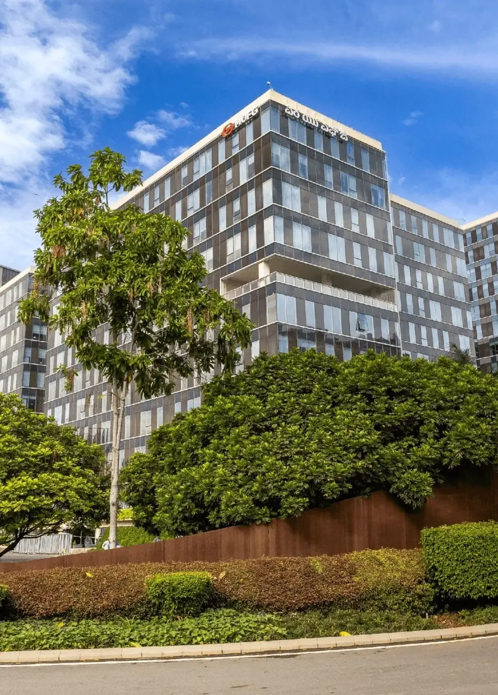 Modern office building with reflective windows behind green trees and bushes under a blue sky.