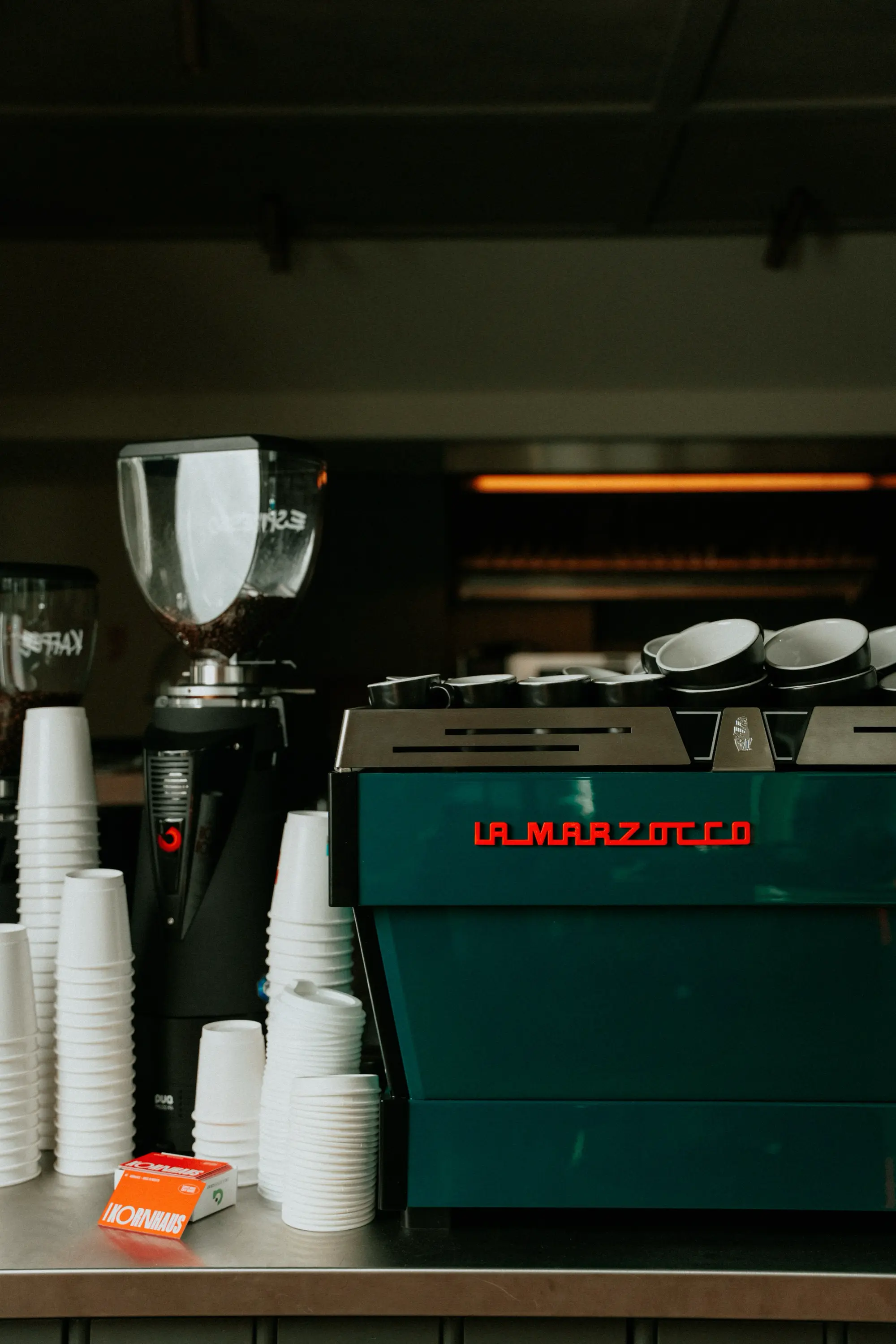 Green La Marzocco espresso machine with stacked coffee cups and a coffee grinder on a counter.