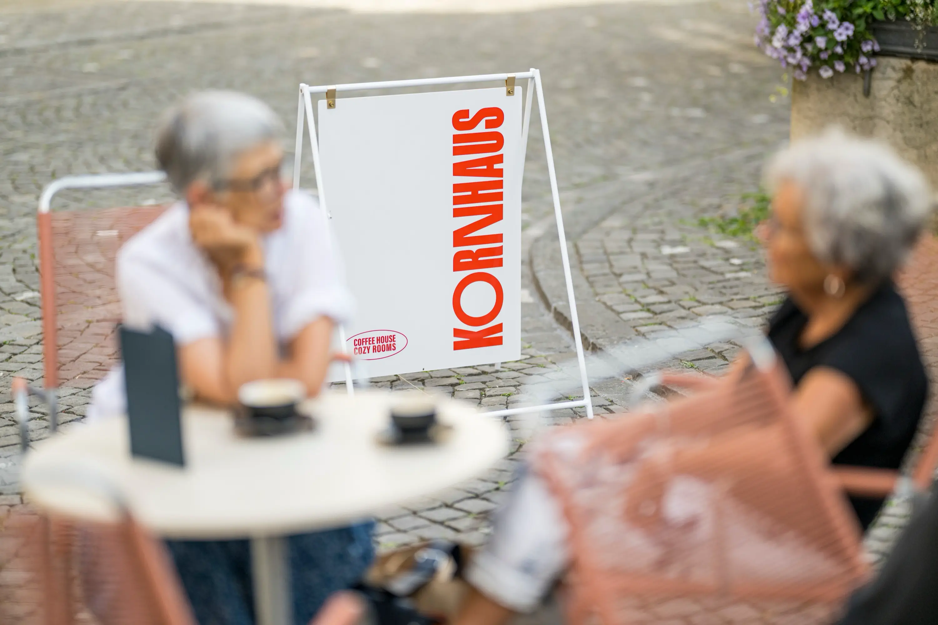 Outdoor café scene with two elderly women sitting at a table and a white sign reading 'KORNHAUS' and 'COFFEE HOUSE COZY ROOMS' in red letters on a cobblestone street.
