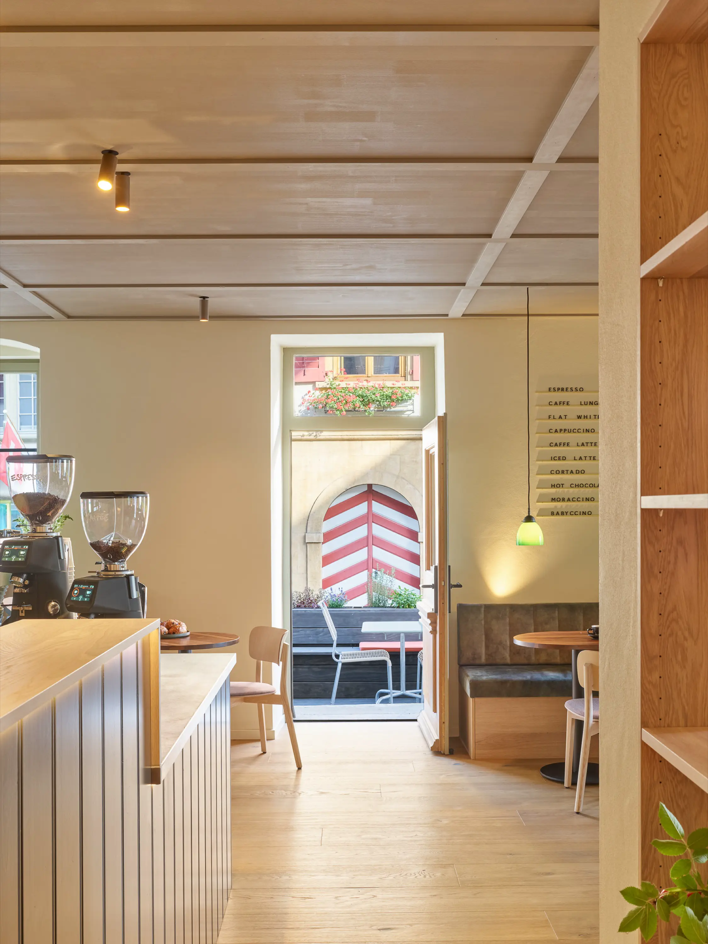 Cozy cafe interior with wooden flooring, coffee grinders on the counter, seating area, and open door leading to outdoor patio with striped red and white wall.