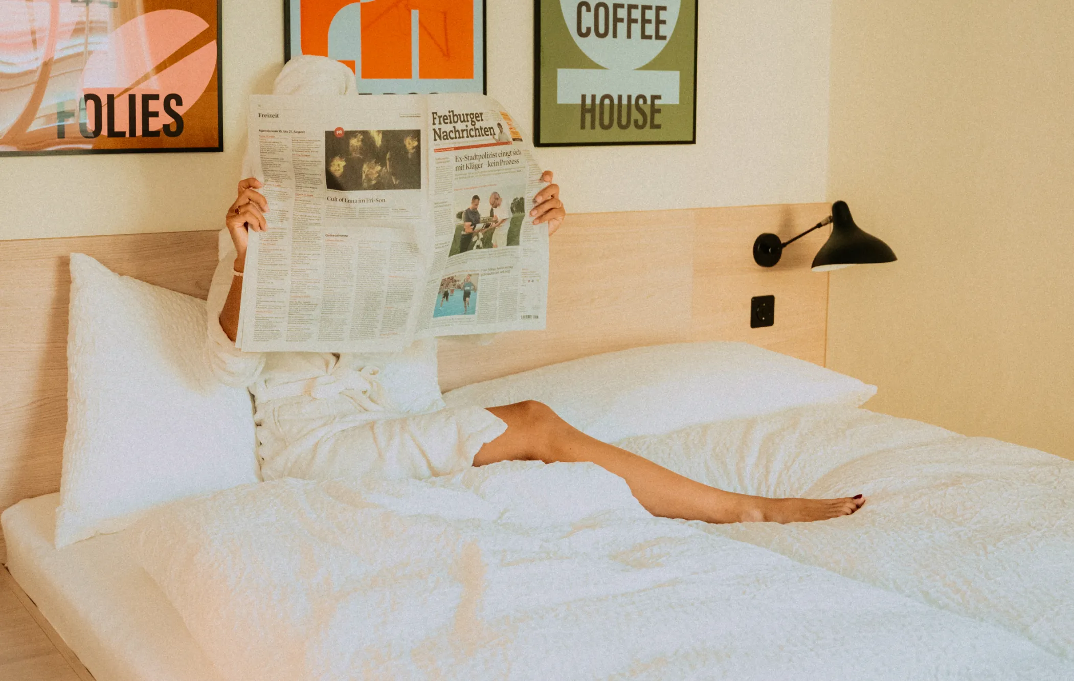 Person in a white robe lounging on a white bed while reading a newspaper in a room with framed wall art and a black wall lamp.