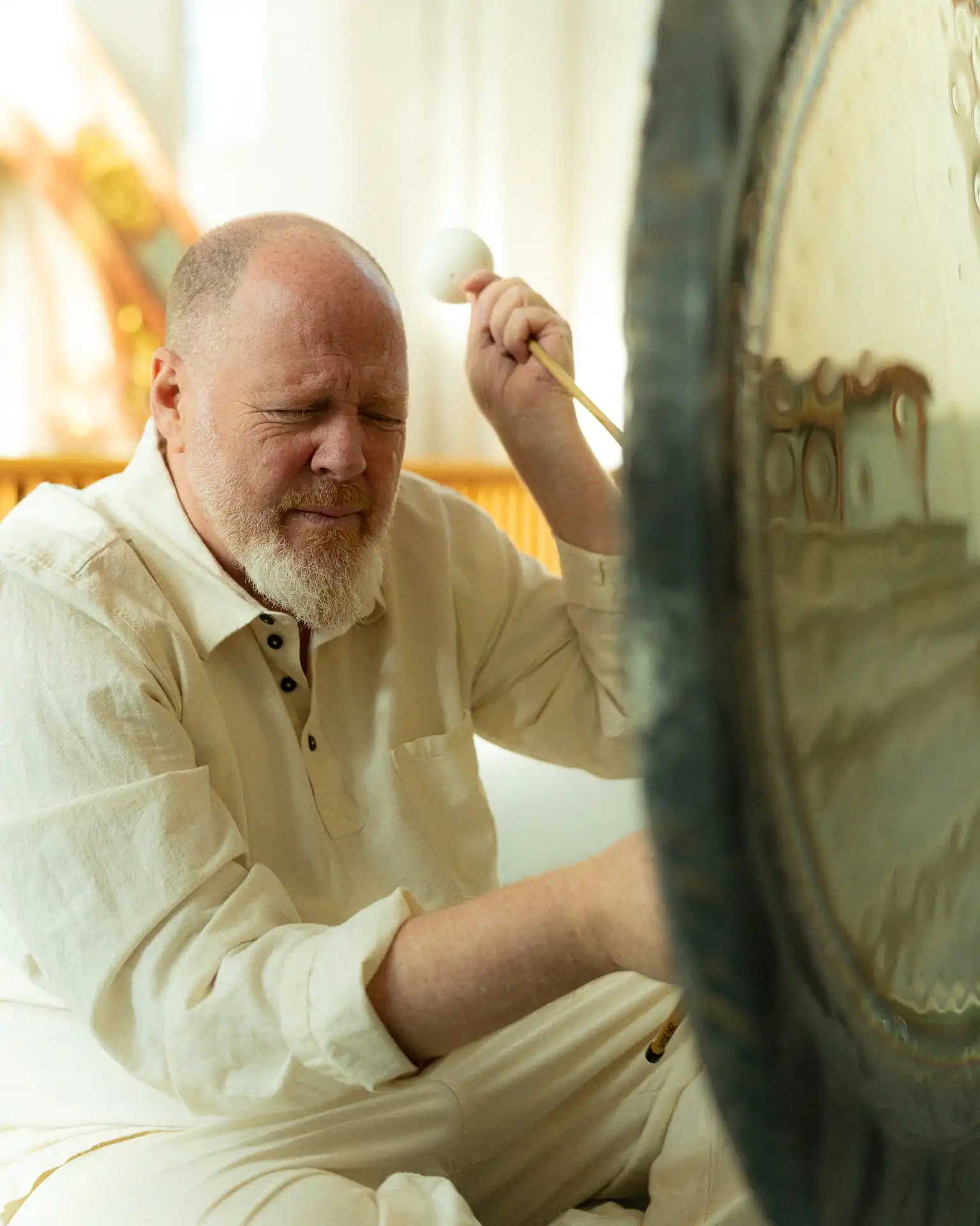 A man with a beard and closed eyes is playing a large gong with a padded mallet in a softly lit room.