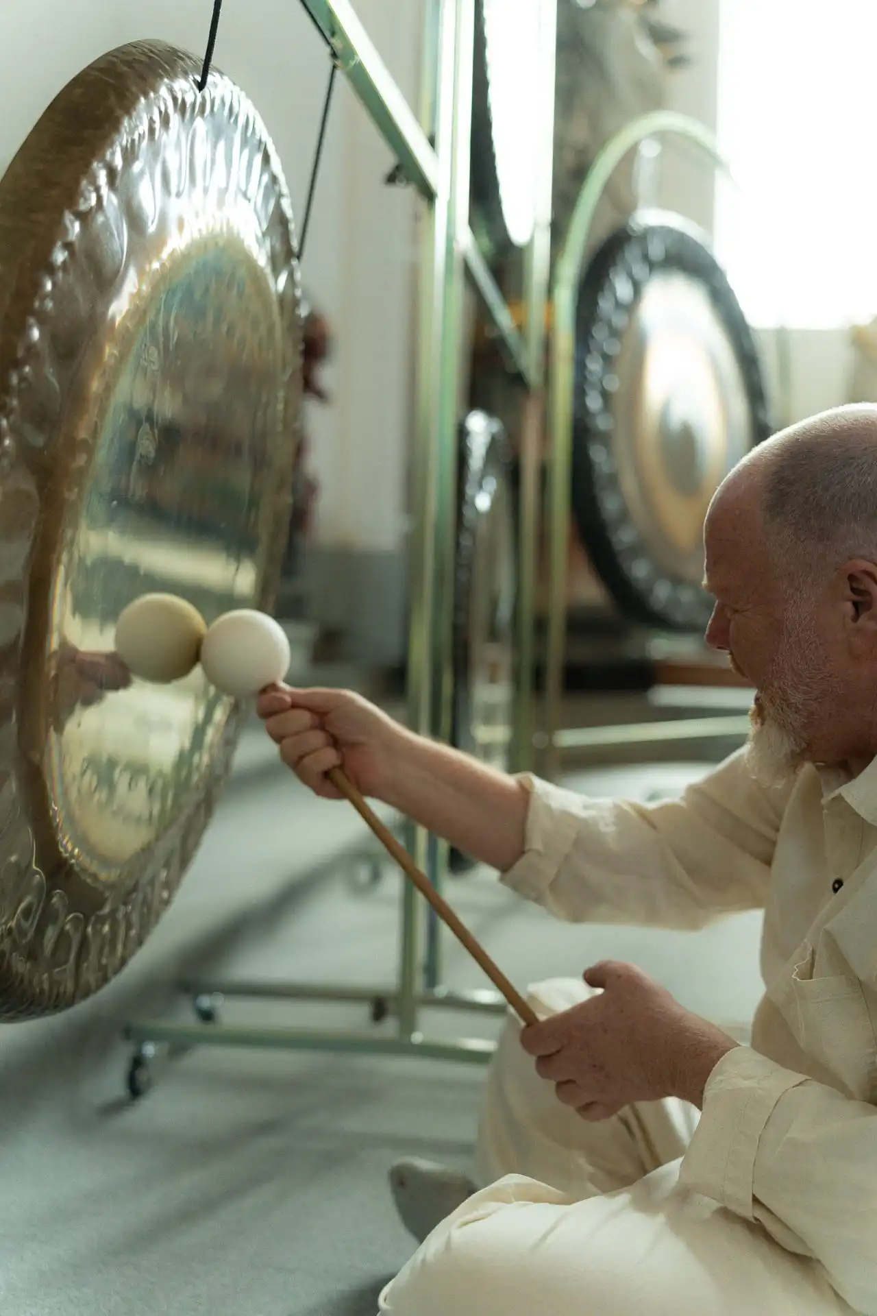 Man with a white beard playing a large gong using two padded mallets in a softly lit room.